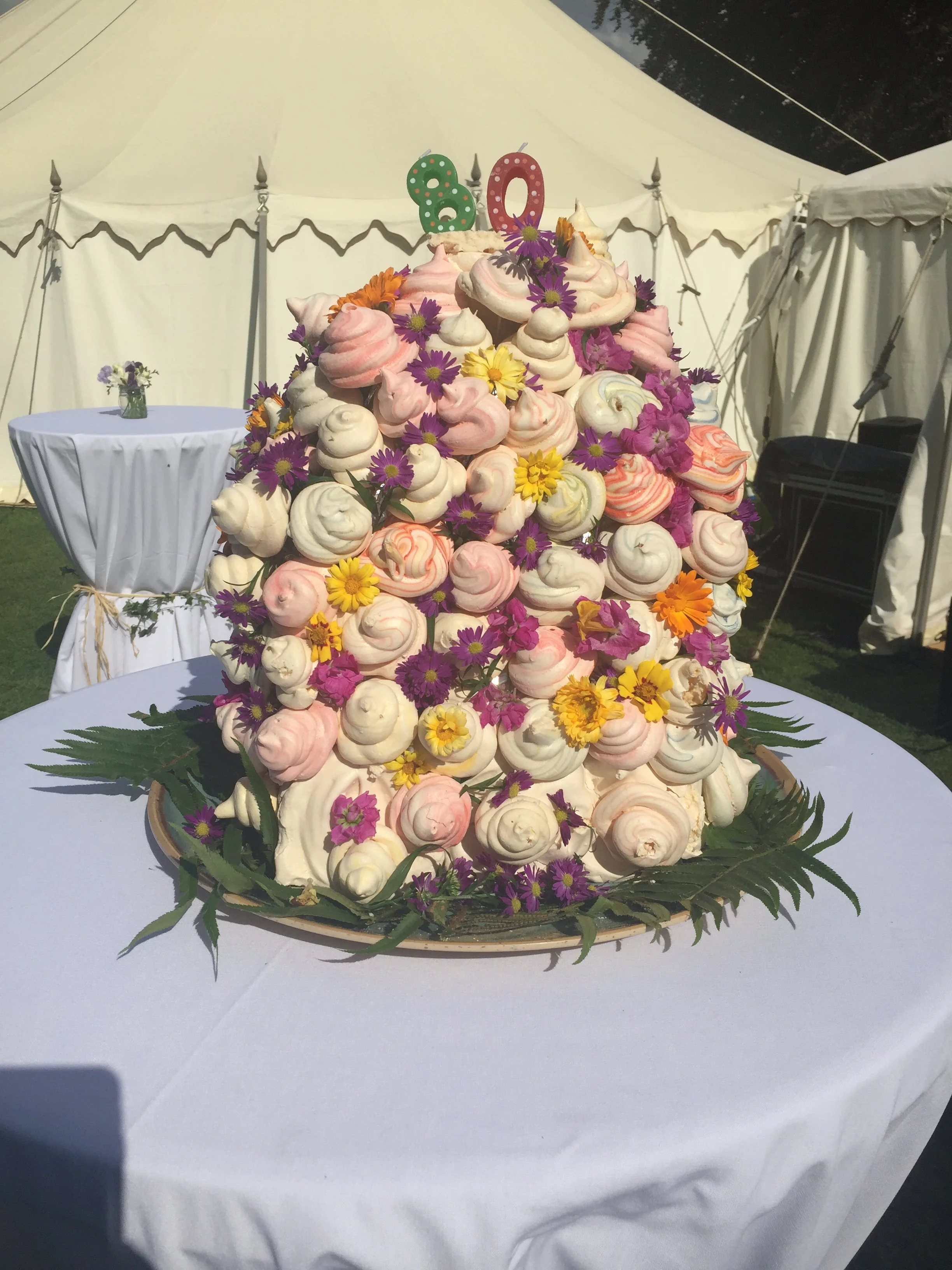 A large cone-shaped dessert tower made of pink and white meringues, decorated with purple, yellow, and orange flowers, with a colorful "80" candle on top, on a white table at an outdoor event.