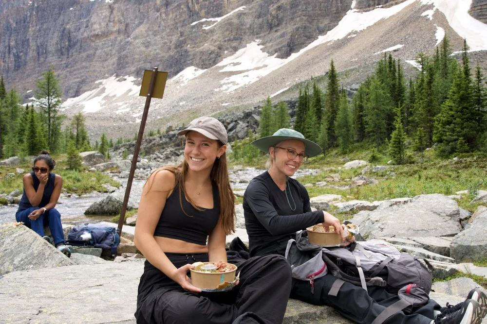 A mother and daughter smiling over a packed trail lunch beside a creek, Lake O'Hara area, Yoho National Park
