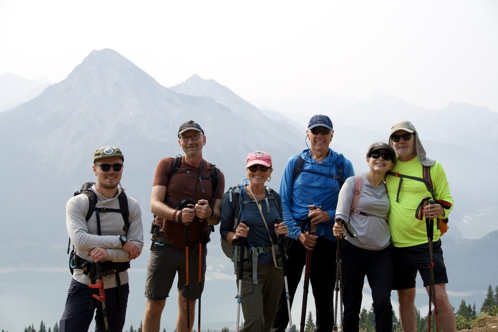 Six hikers with trekking poles smiling together on a ridge above a lake in the Canadian Rockies