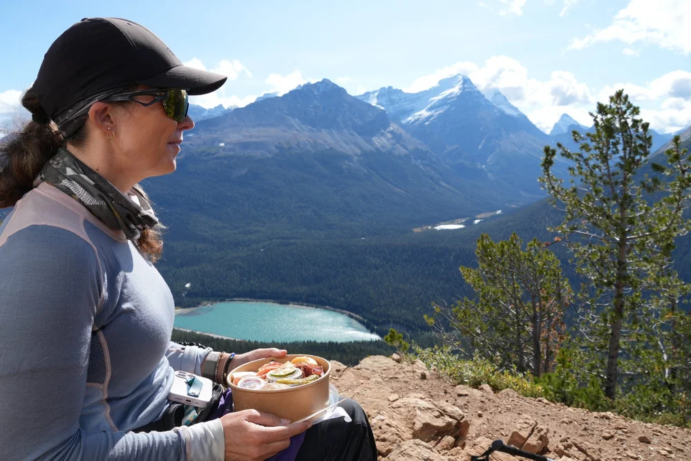 A hiker holding a trail lunch bowl at Paget Lookout above a turquoise lake, Yoho National Park