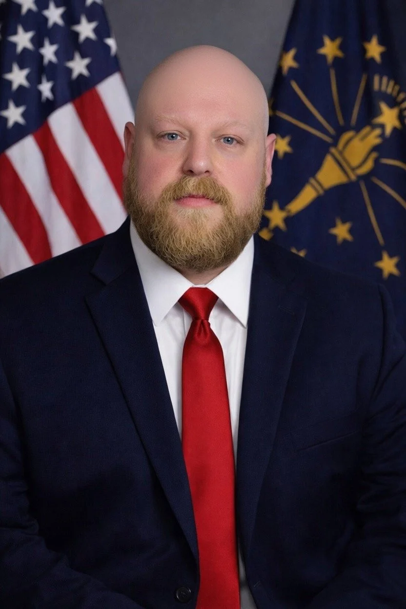 A man with a bald head and beard wearing a dark suit, white shirt, and red tie, in front of American flags and Indiana state flag.
