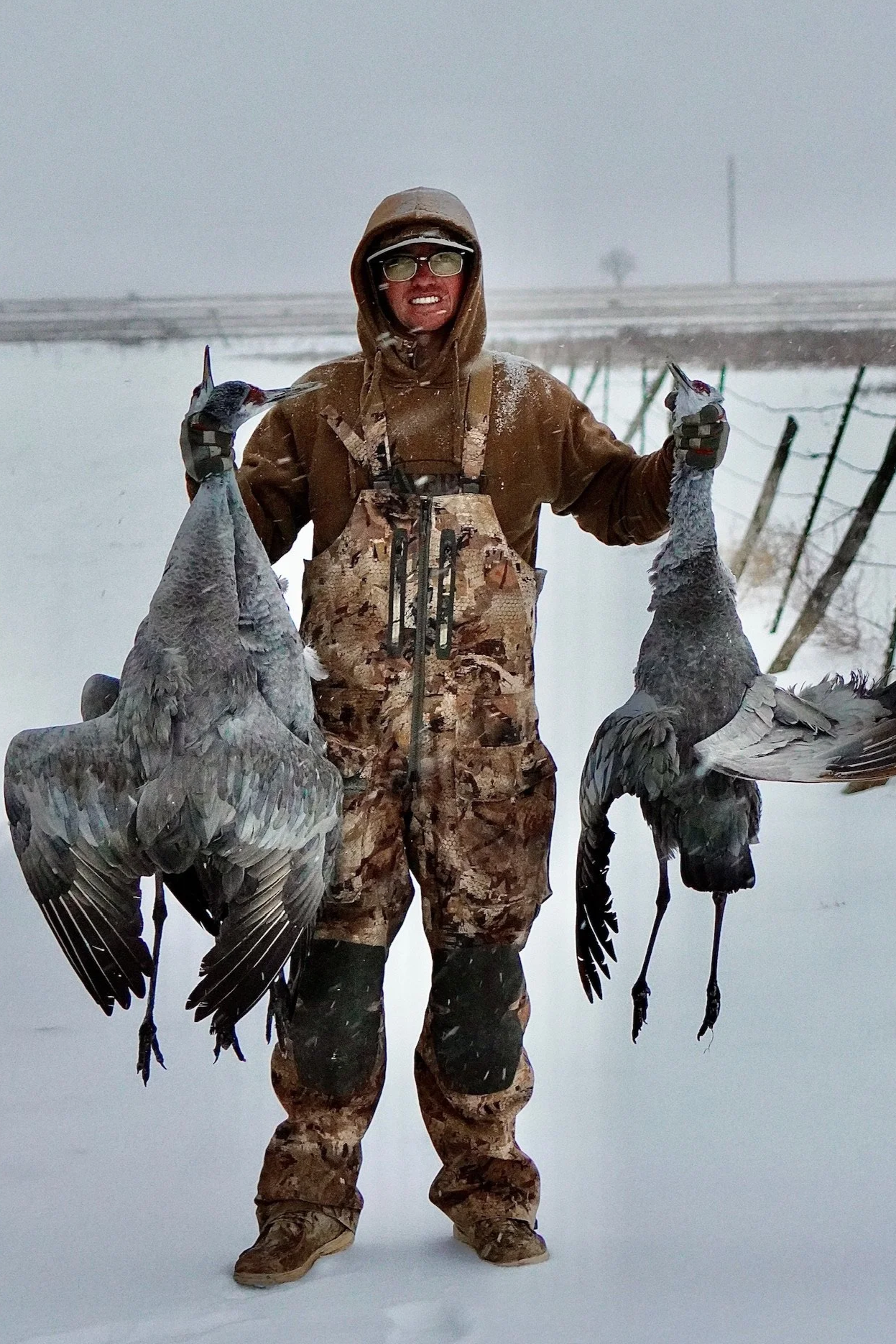 A person dressed in camouflage and winter clothing holding three sandhill crane in a snowy landscape, likely after a hunting trip.