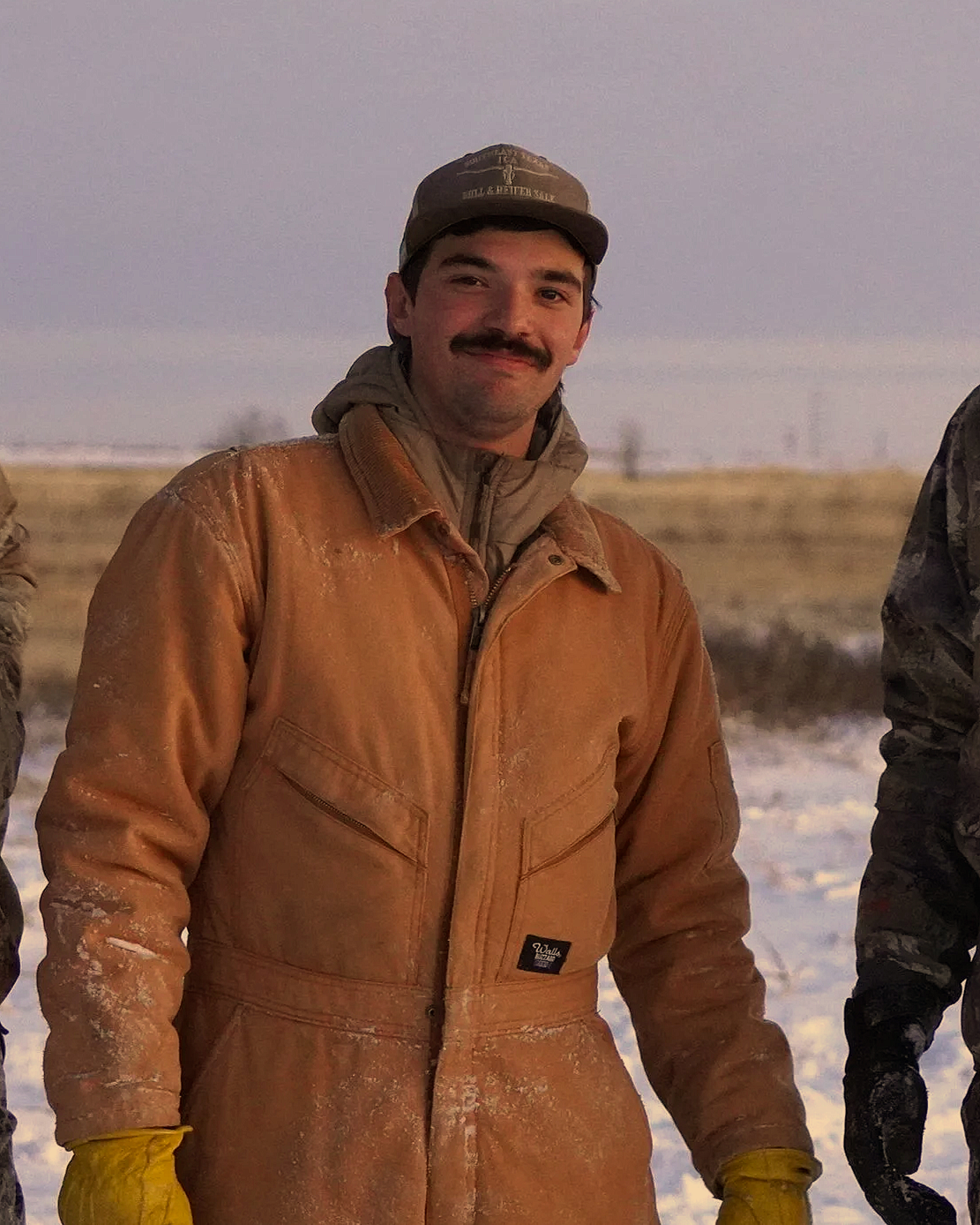 A man smiling outdoors in a snowy field, wearing a brown leather jacket, a gray hoodie, a cap, and yellow gloves.