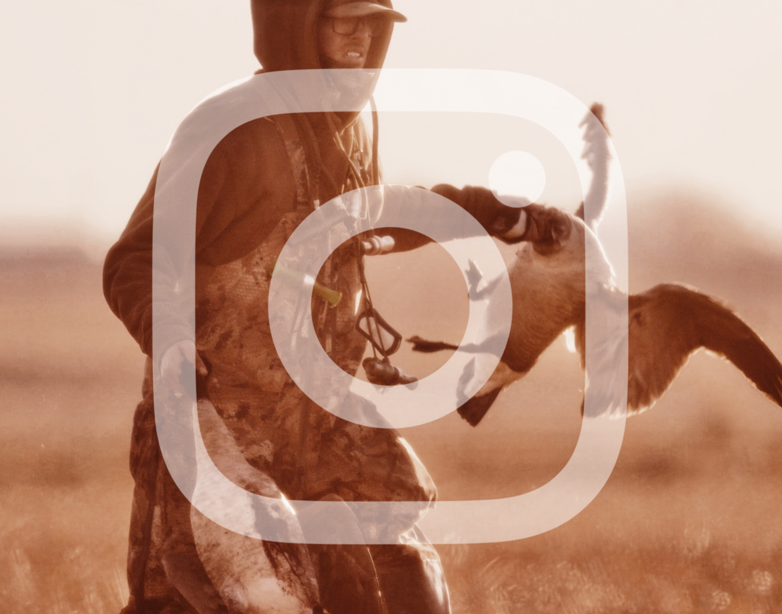 Person dressed in camo, holding a dead canada goose in an open field.