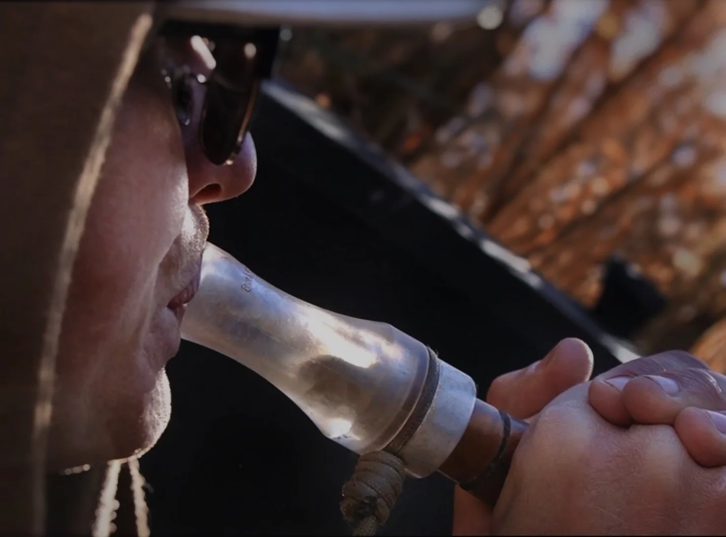 Person wearing sunglasses and hoodie, using a goose call to lure in birds while staying discreet in an A-frame hunting blind.