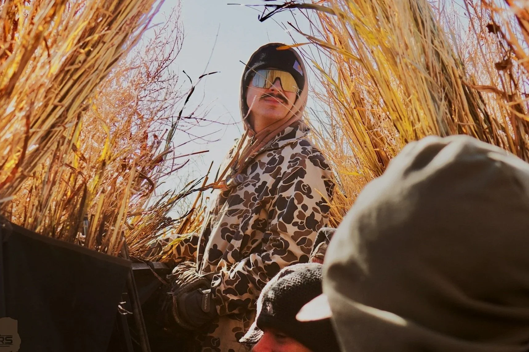 Person in camouflage jacket and sunglasses in a wheat field, looking up.