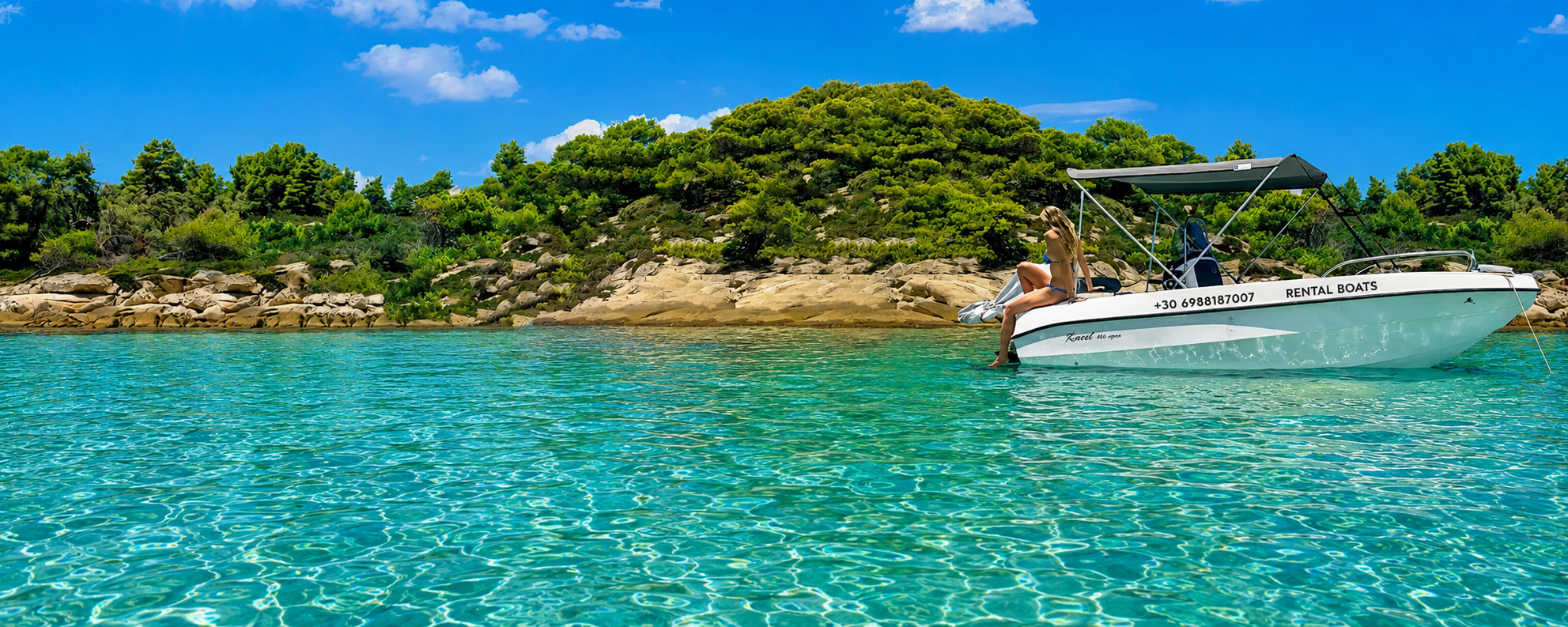 Woman sitting on the edge of a white boat in clear turquoise water near a rocky and green hillside on a bright sunny day.