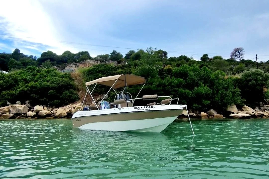 A small motorboat named Blue Pearl anchored near a rocky shoreline with dense green bushes and trees, under a partly cloudy sky.