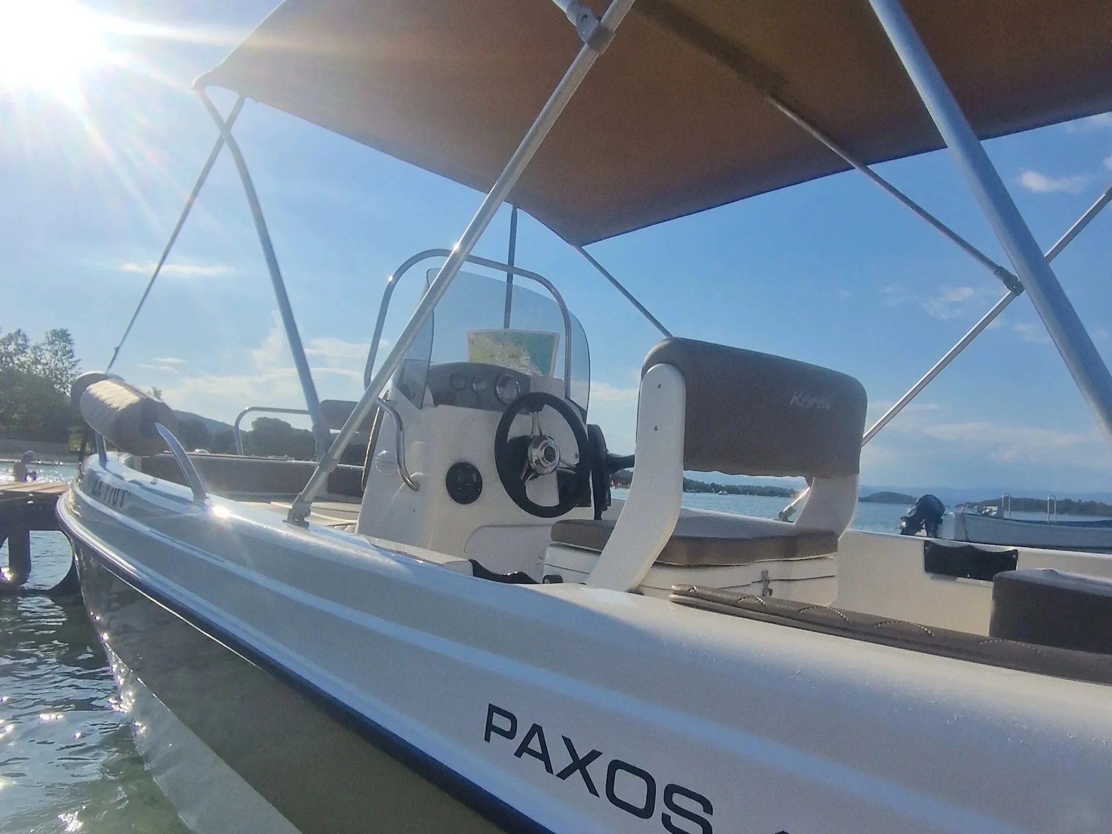 A white motorboat docked in a calm water body with blue sky and some clouds, with a name starting with 'PAXOS' visible on the side and part of the boat's interior including the steering wheel and dashboard.