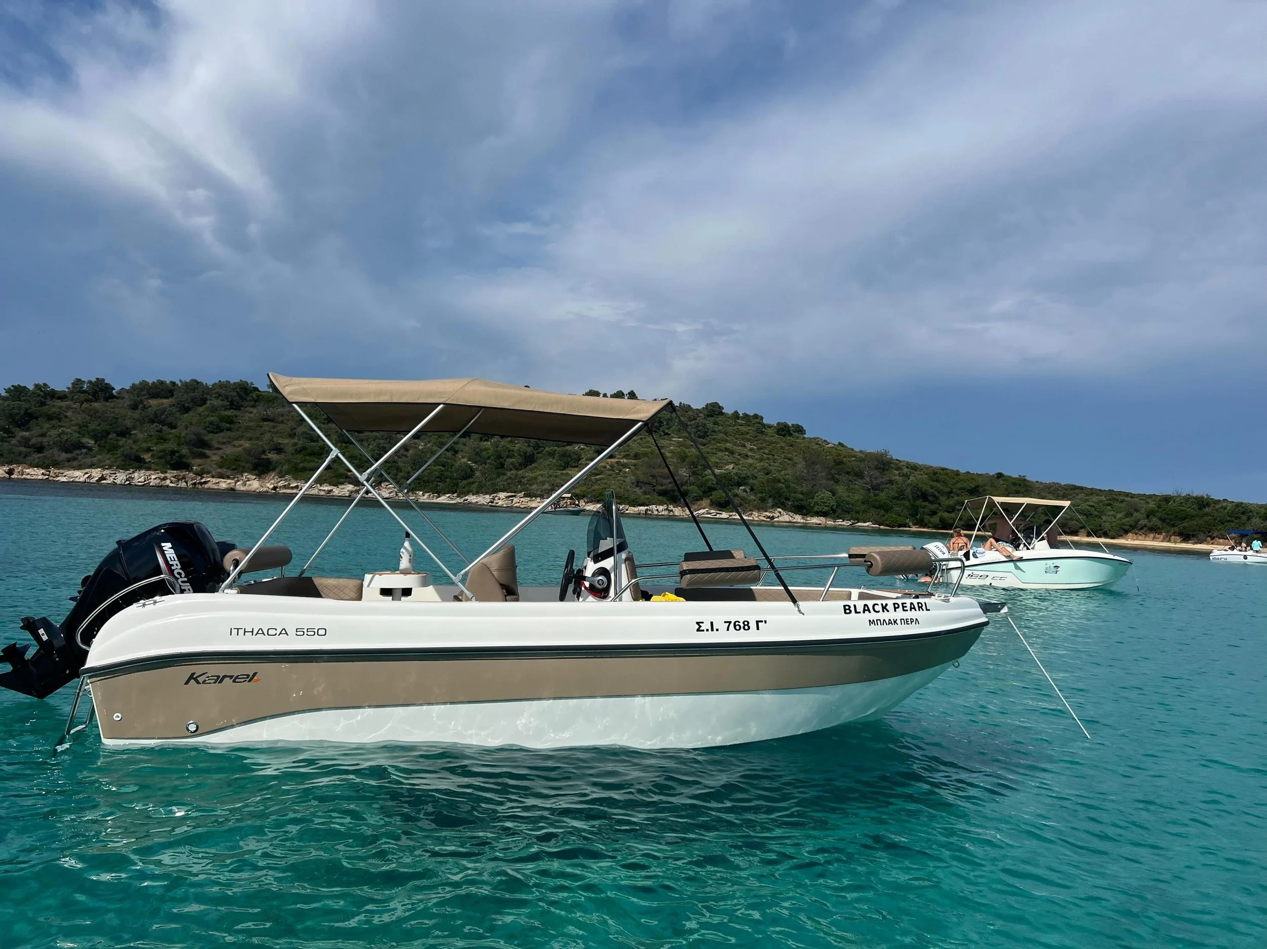 A white motorboat named 'Black Pearl' floating on turquoise water near a green hillside, with a canopy and outboard motor, and a clear sky with some clouds.