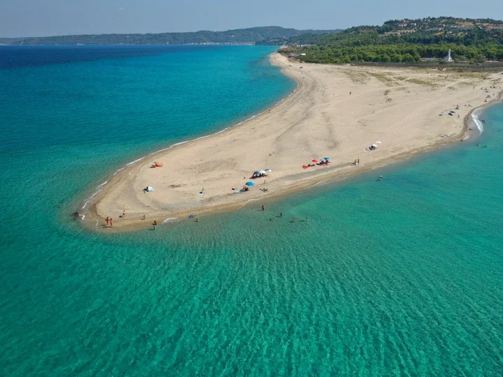 Aerial view of a sandy beach peninsula surrounded by turquoise water with people, umbrellas, and boats, and a green hillside in the background.