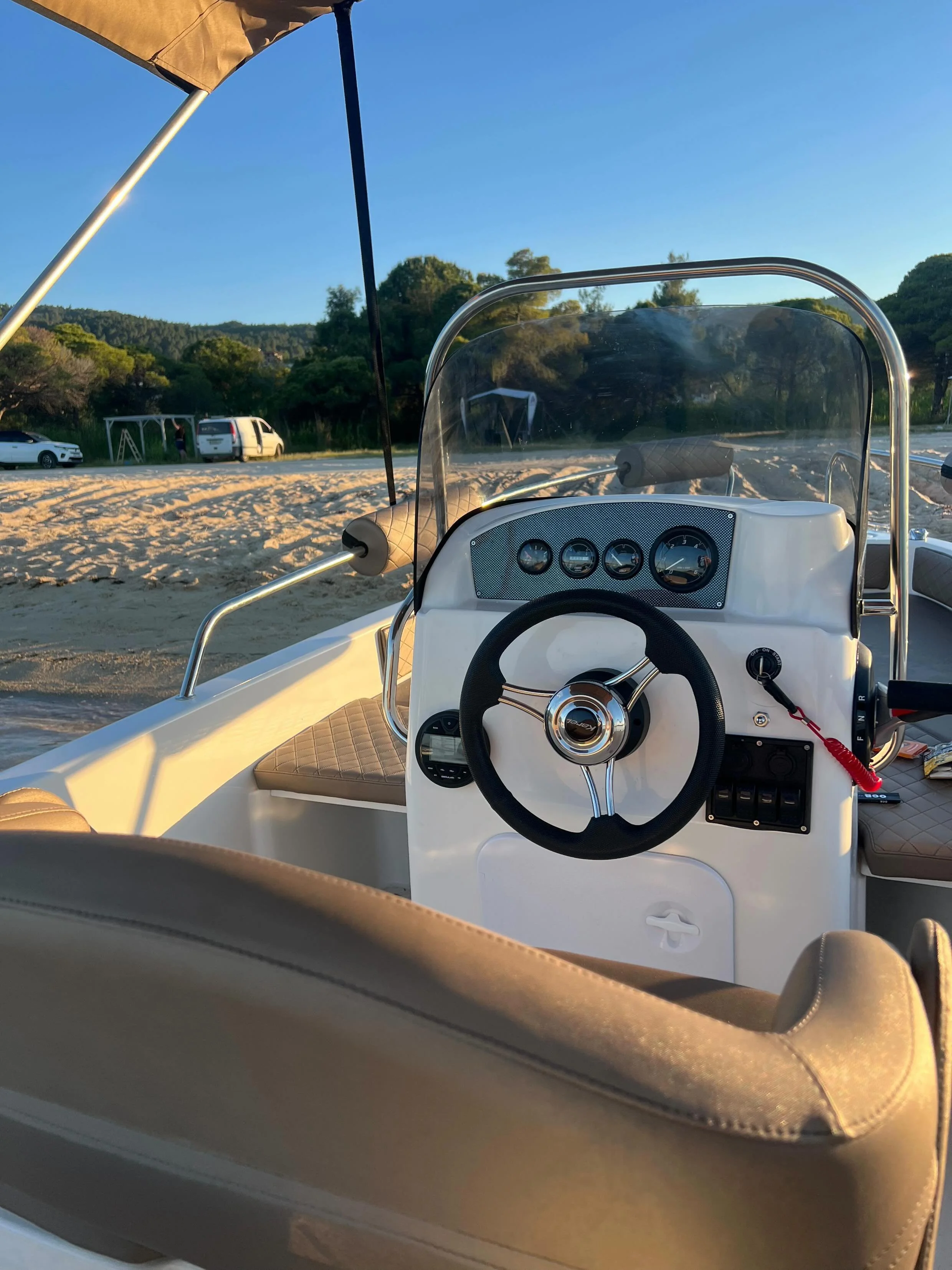 View from inside a boat showing the dashboard with gauges, a windshield, and part of a steering wheel, with a sandy beach, parked cars, and green trees in the background under a clear blue sky.
