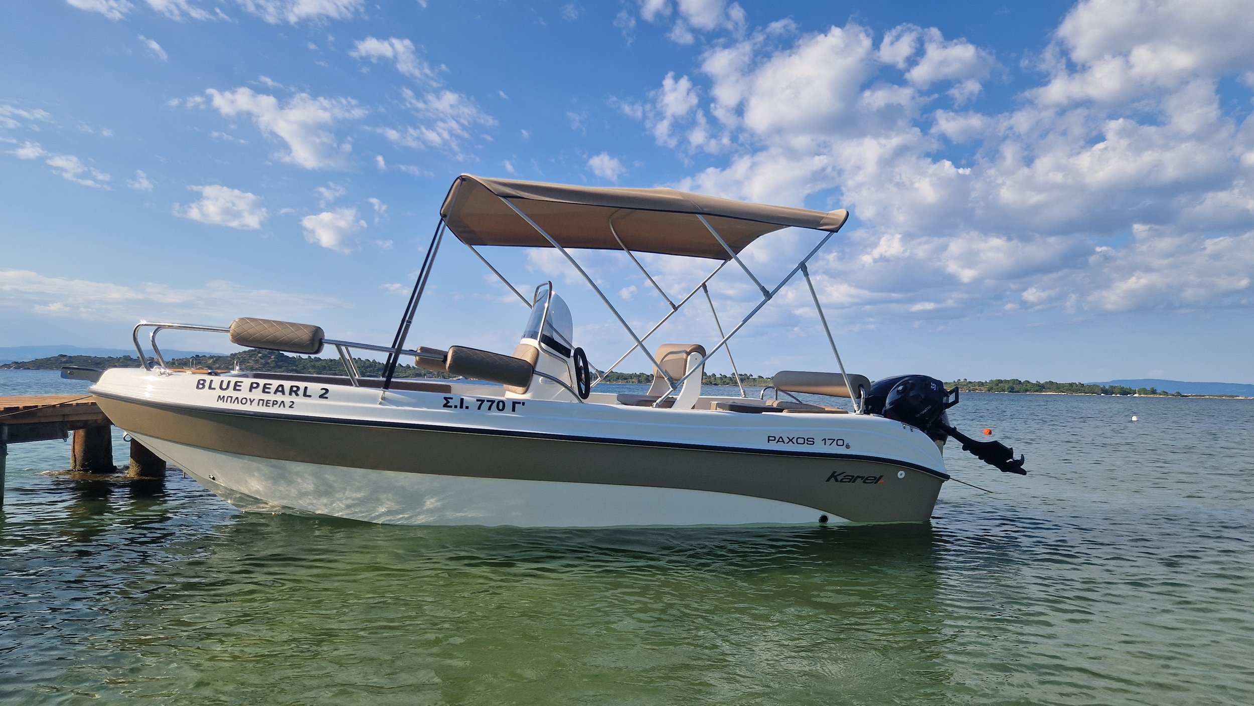 A small white boat named BLUE PEARL 2 with a canopy, moored near a wooden pier on calm water against a blue sky with scattered clouds.