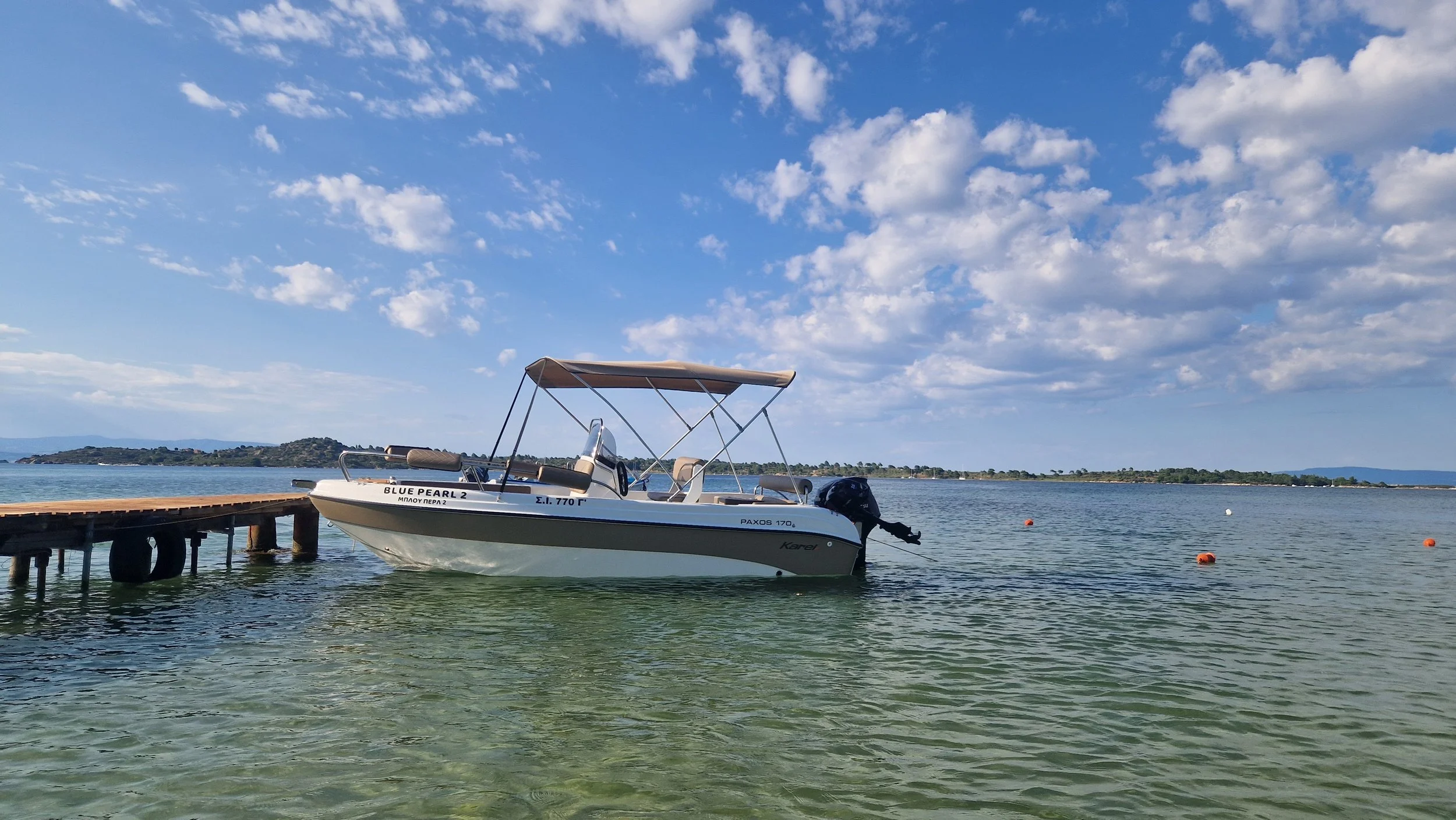 A small white motorboat docked at a wooden pier on a calm lake, with a distant shoreline and a partly cloudy blue sky in the background.