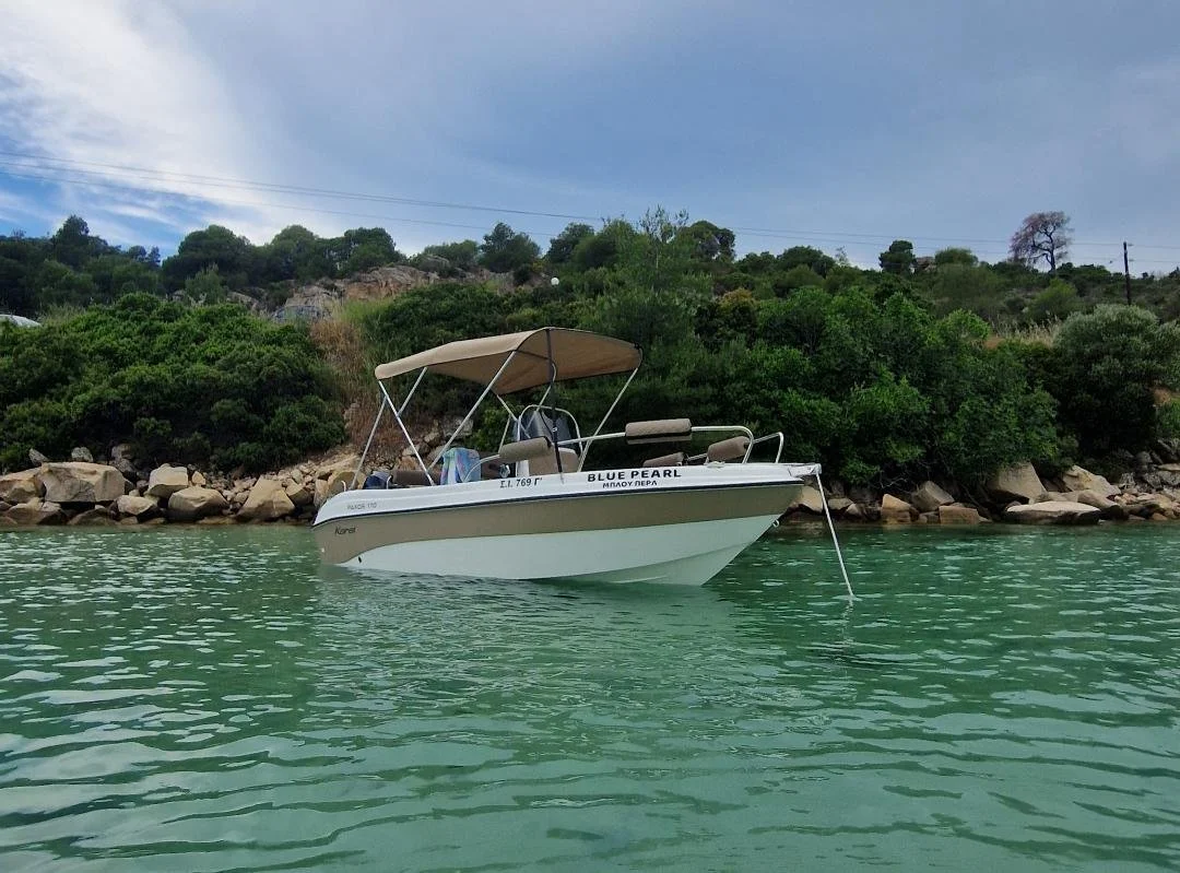 A small white motorboat named 'Blue Pearl' is anchored near a rocky and wooded shoreline on calm, greenish water with an overcast sky overhead.
