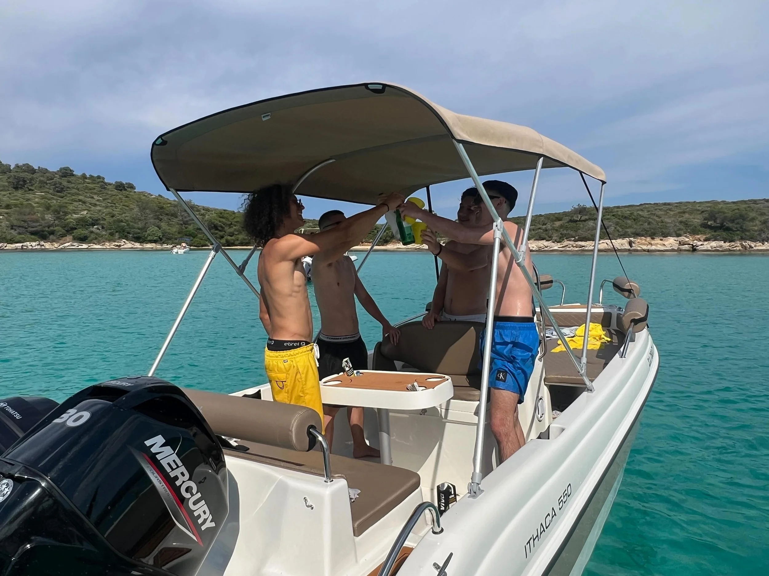 Three shirtless men on a boat sharing drinks and smiling, with a body of water and green hillside in the background.