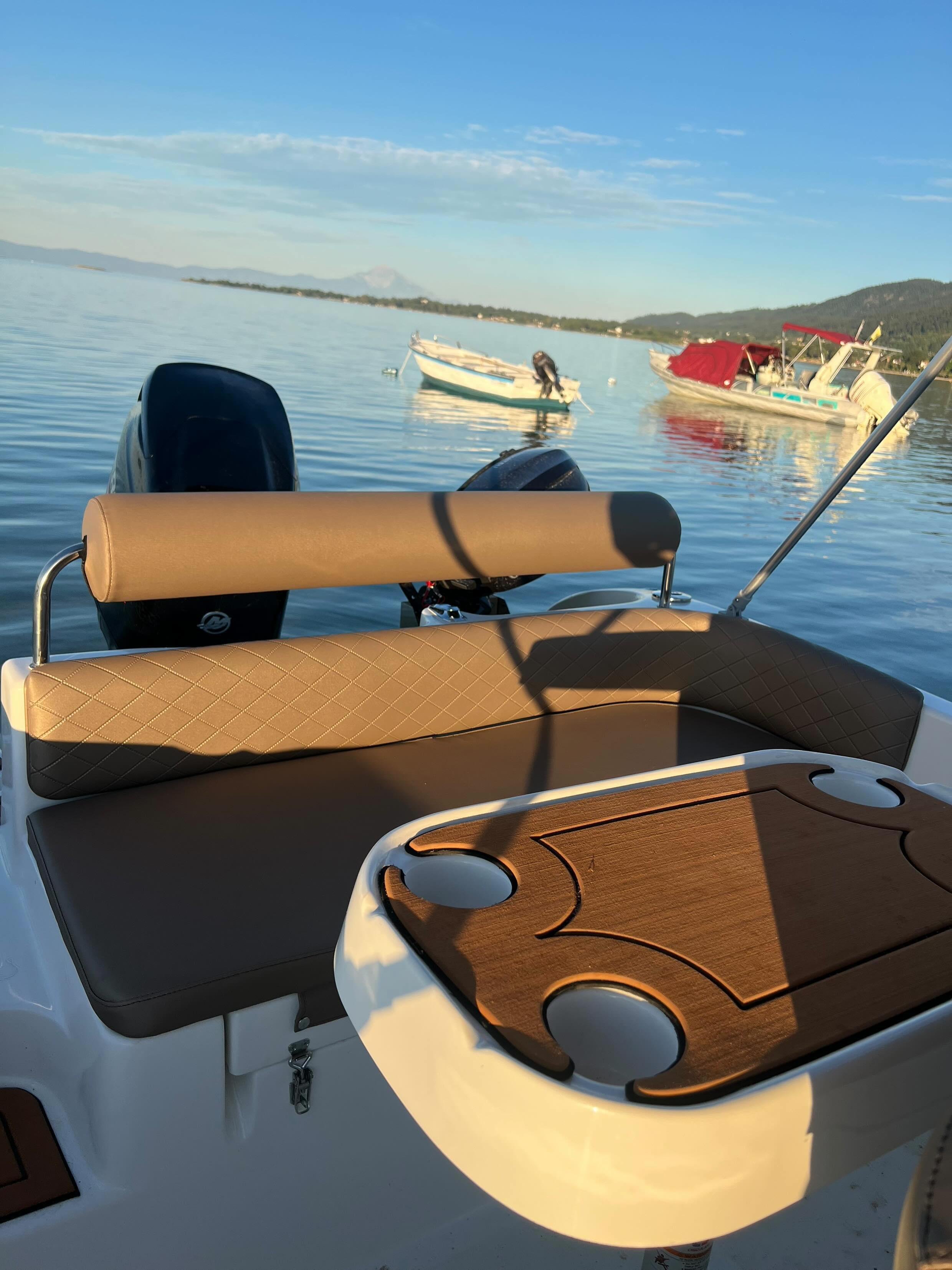 The image shows the back part of a boat with a cushioned seating area and a wood-like table with a curved design, situated on calm water with two boats and a person near a small inflatable boat in the background, under a clear sky.