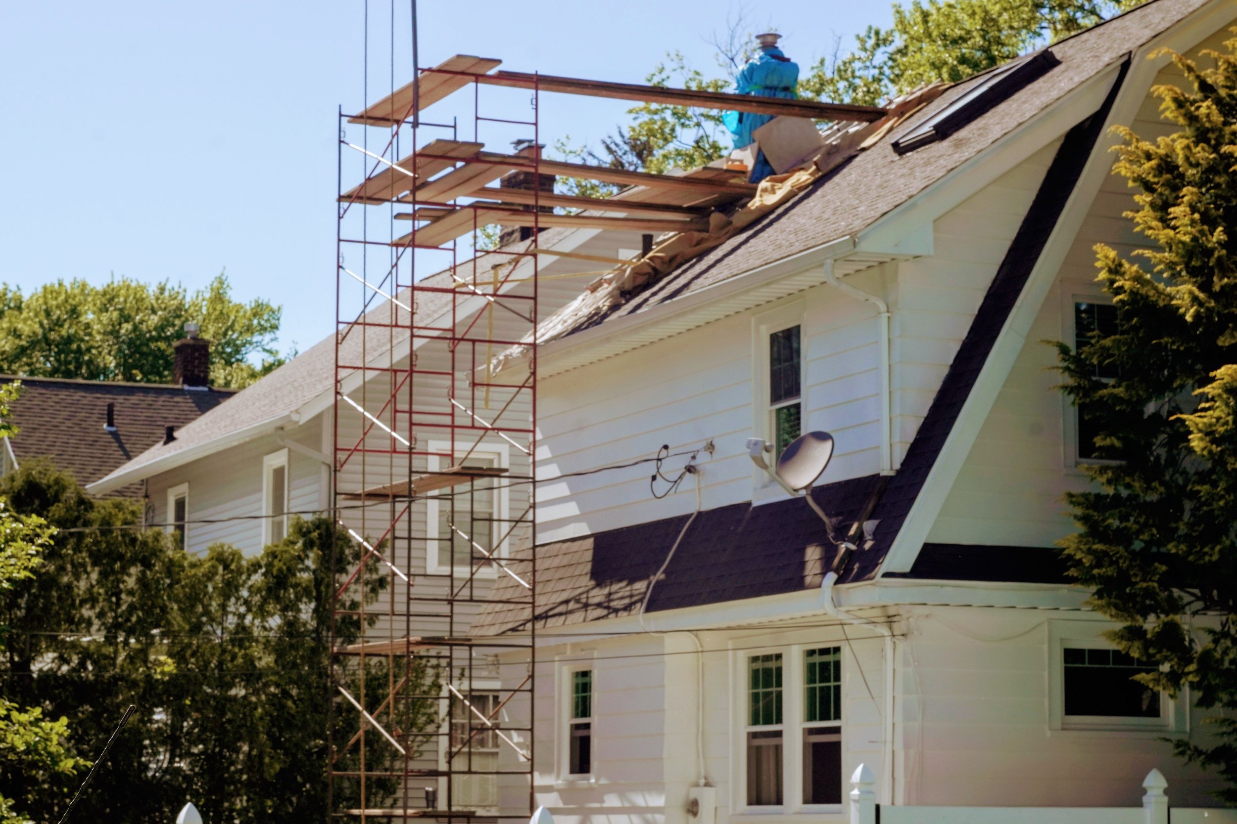 A house undergoing roof repairs with scaffolding set up on its side. A worker is on the roof, which is partially covered with brown protective cloth. The house is white with multiple windows, and trees are visible around it.