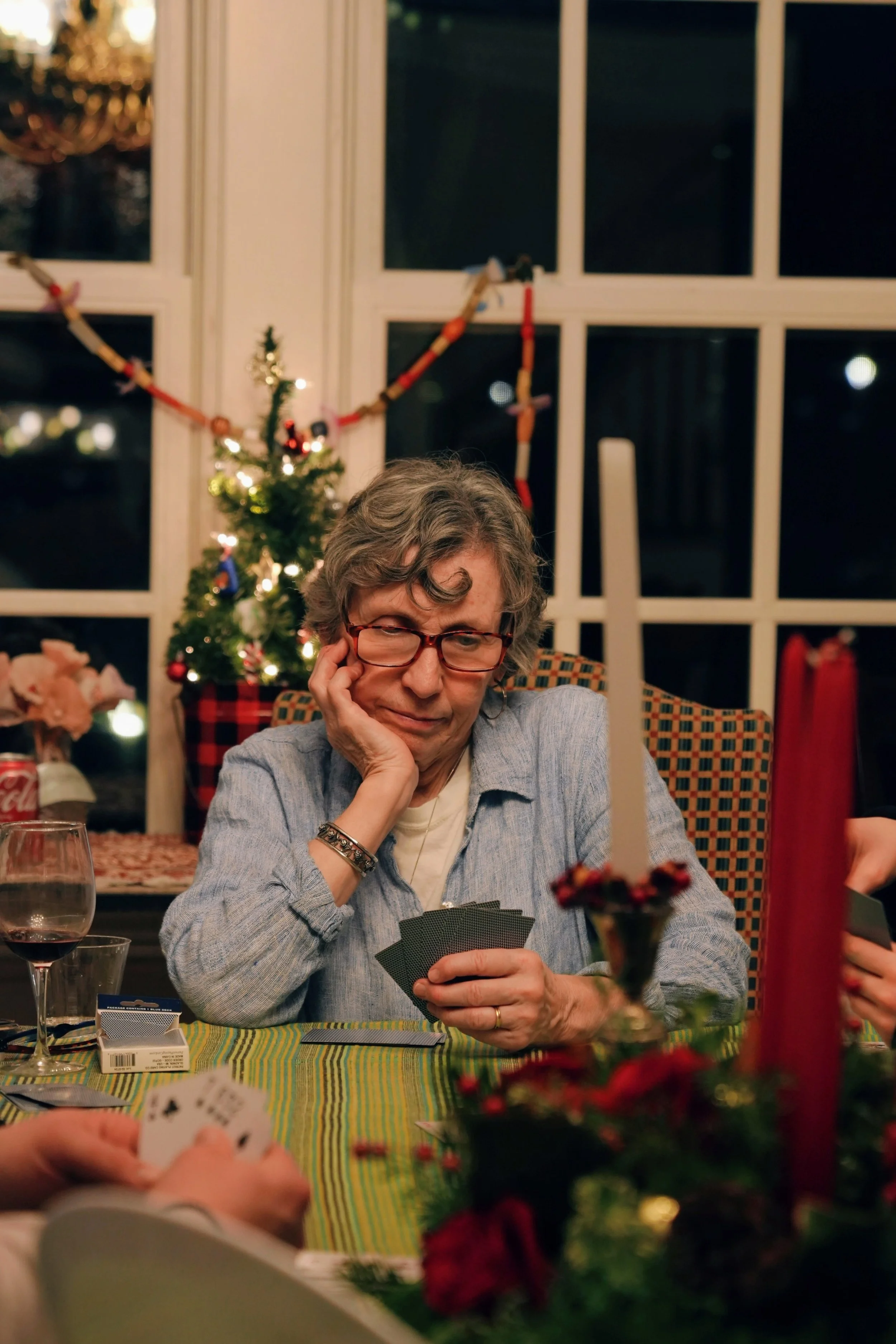 An older woman with glasses, gray hair, and a gray shirt is sitting at a table playing cards, with a thoughtful expression. The table has a festive tablecloth, a glass of wine, and holiday decorations, including a small decorated Christmas tree in th