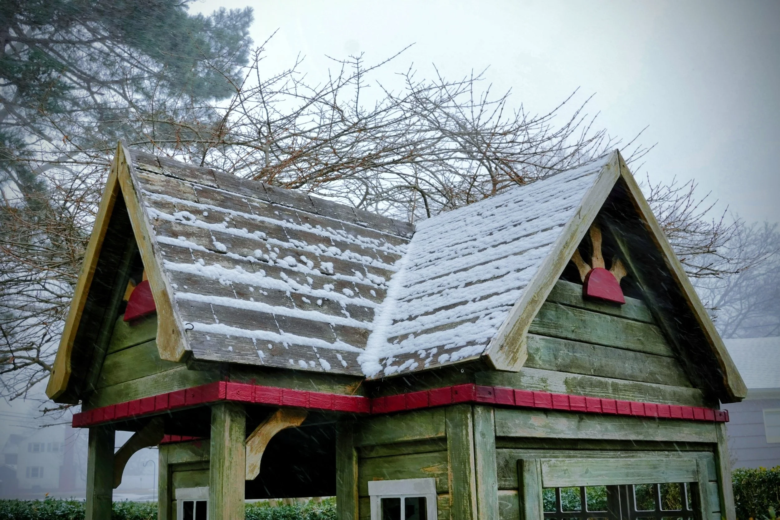 A small wooden playhouse with a snow-covered roof and leafless trees in the background.