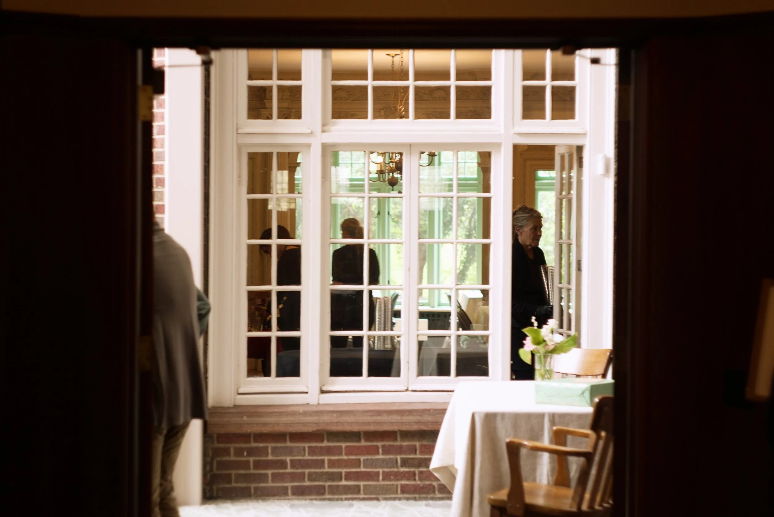 View of a dining room through a window, with a table, chairs, and a vase of flowers. Several people are seen inside, including an older woman standing near the window. The scene is framed by dark edges of a doorway.