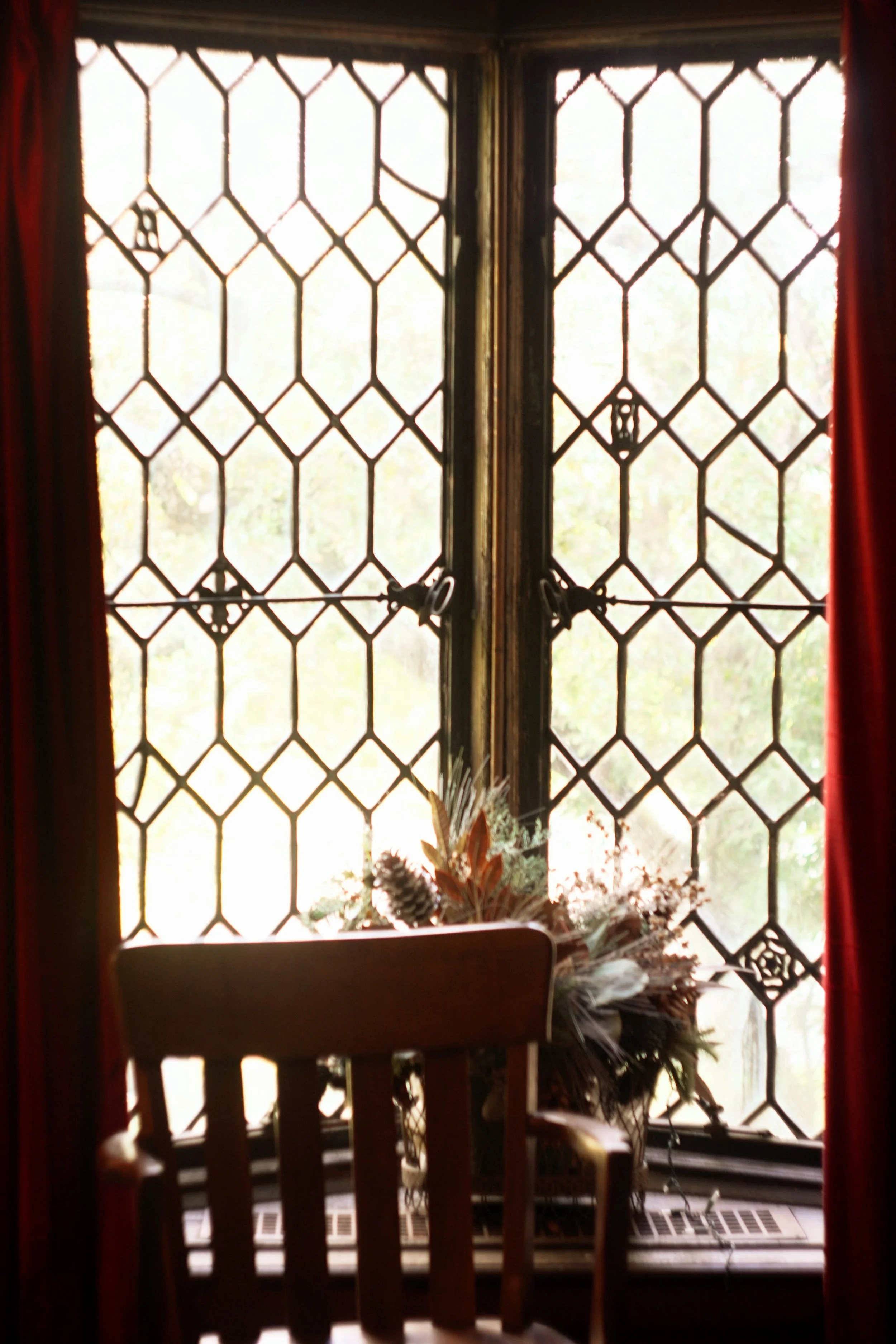 A window with diamond-shaped glass panes and black frames, partially open with round window latches, with a wooden chair and a bouquet of dried flowers and pinecones on the windowsill. Red curtains are on either side of the window.