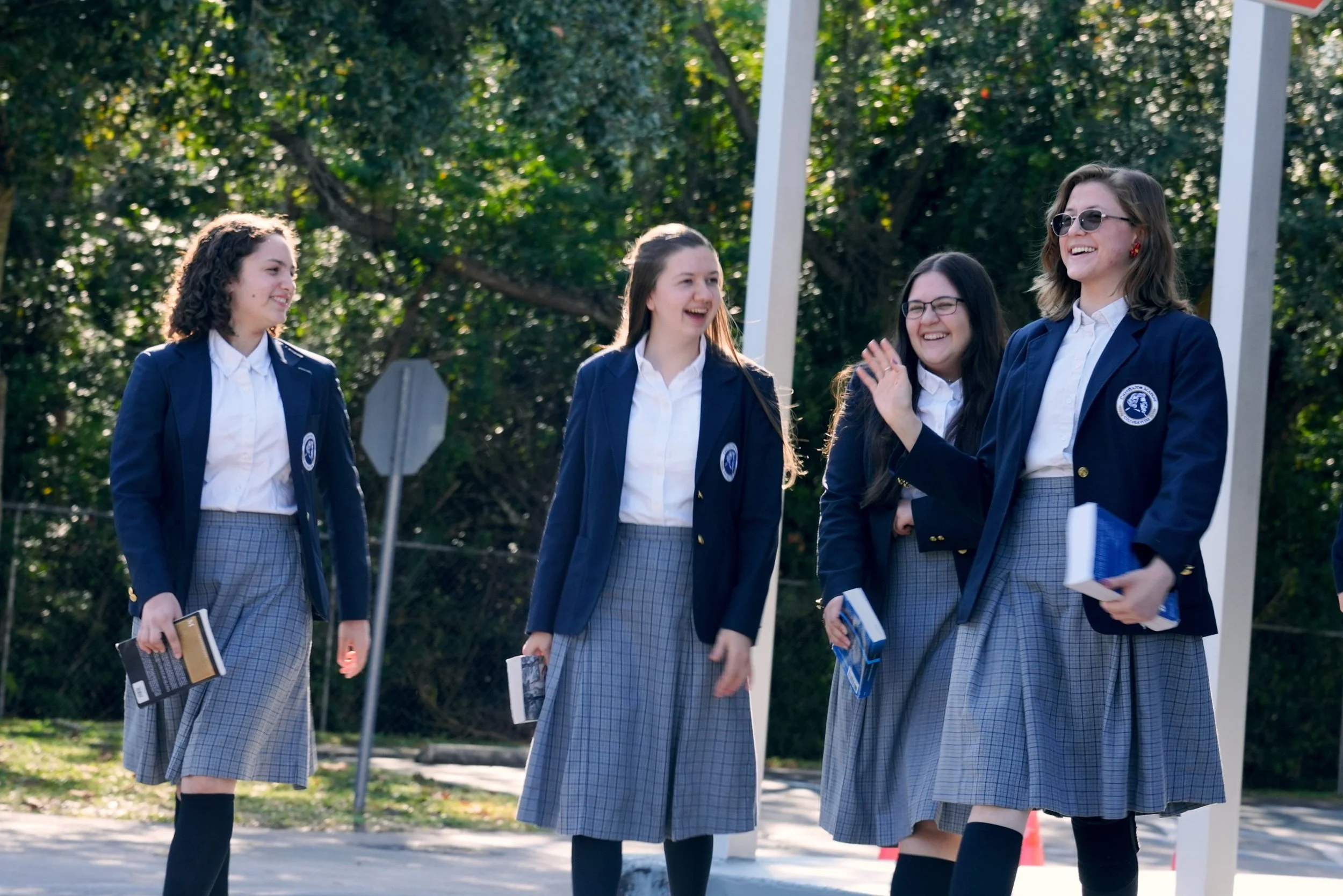 Four teenage girls in school uniforms laughing and talking outdoors, holding books, with trees in the background.