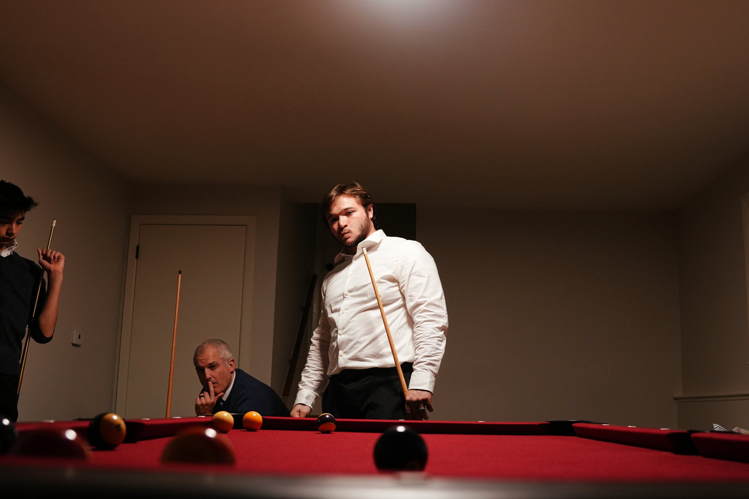 Three men are playing pool in a room with beige walls and a red pool table. The man in the center, wearing a white shirt, looks directly at the camera while holding a pool cue. The other two men are on the left, one partially visible and holding a cu