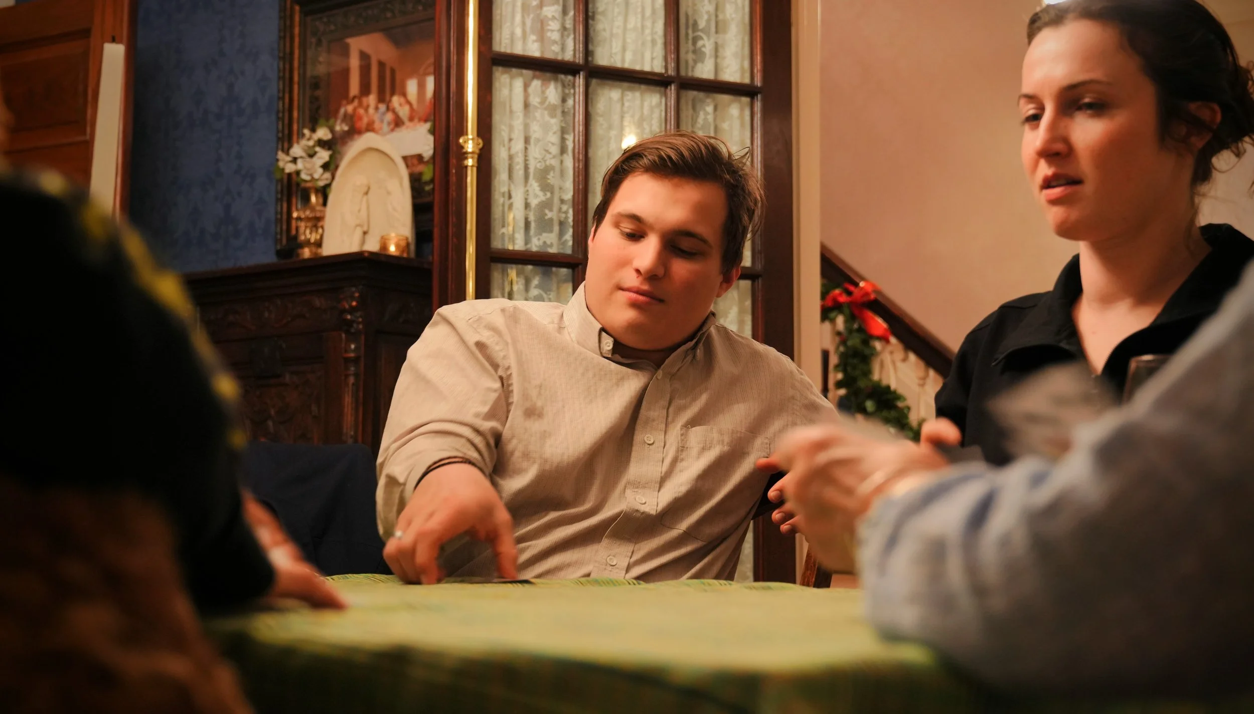 Young man sitting at a table with a woman, in a cozy, decorated interior home setting, possibly during a holiday gathering.