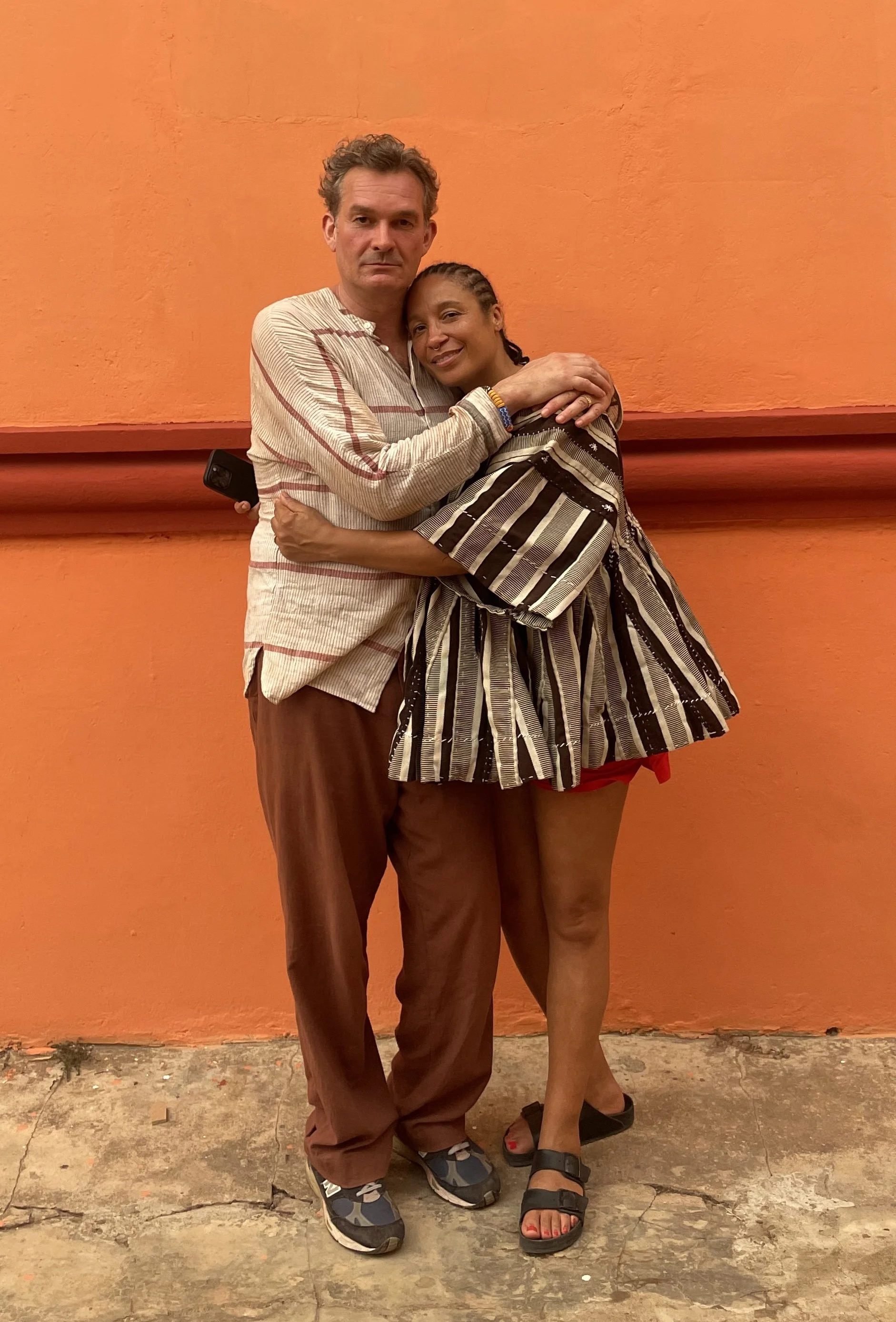 A man and a woman hugging against an orange wall, with the man holding a cellphone behind his back. The man is wearing a beige striped shirt and brown pants, and the woman is dressed in a black and white striped top and red shorts, wearing sandals.