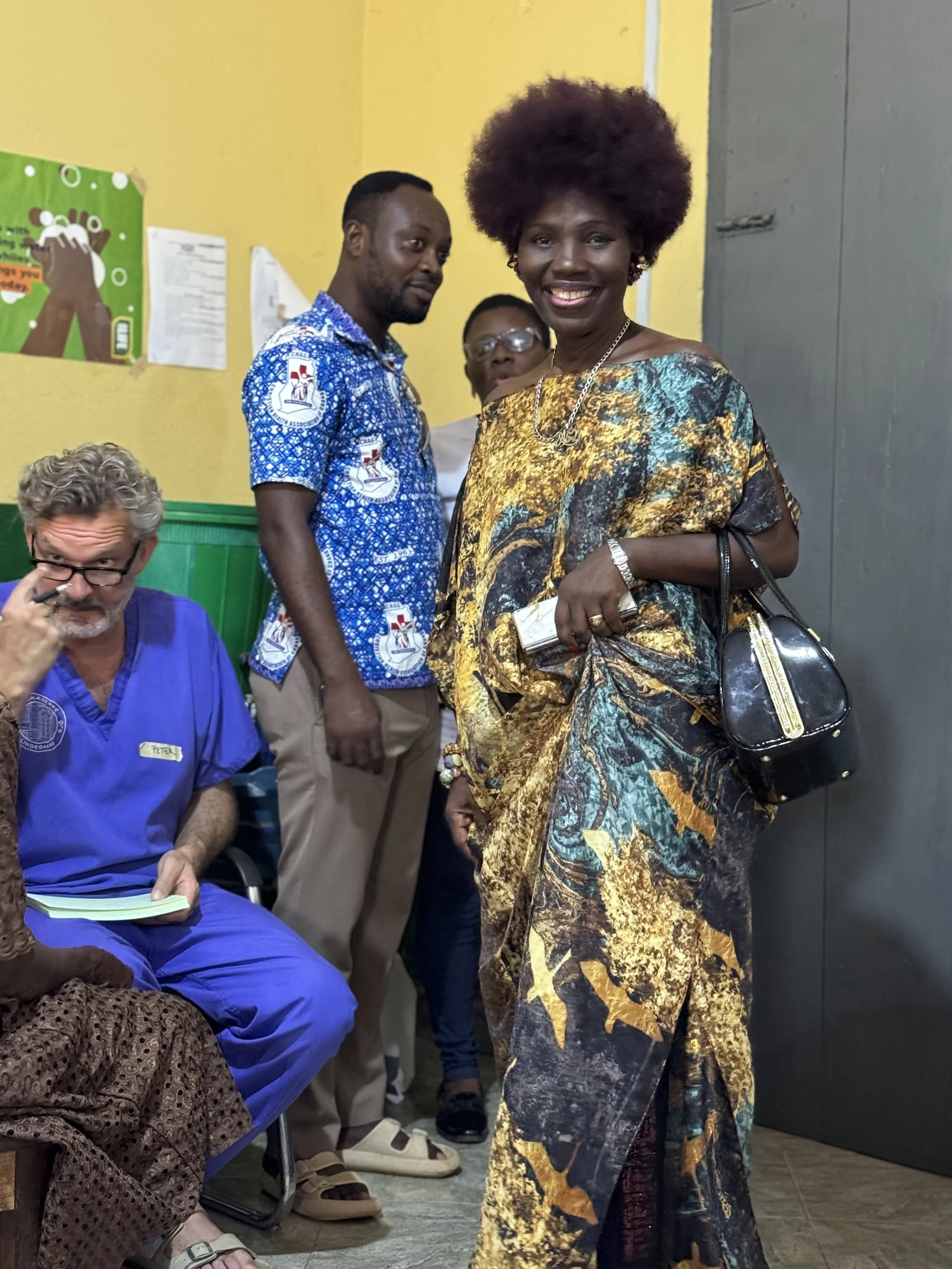 A group of people indoors, with a woman in the foreground wearing a colorful, patterned dress and smiling at the camera. She is holding a black handbag and a small clutch. Other people are visible behind her, including a man in a blue patterned shirt and another person with glasses. The setting appears casual, with yellow walls and some posters on the wall.