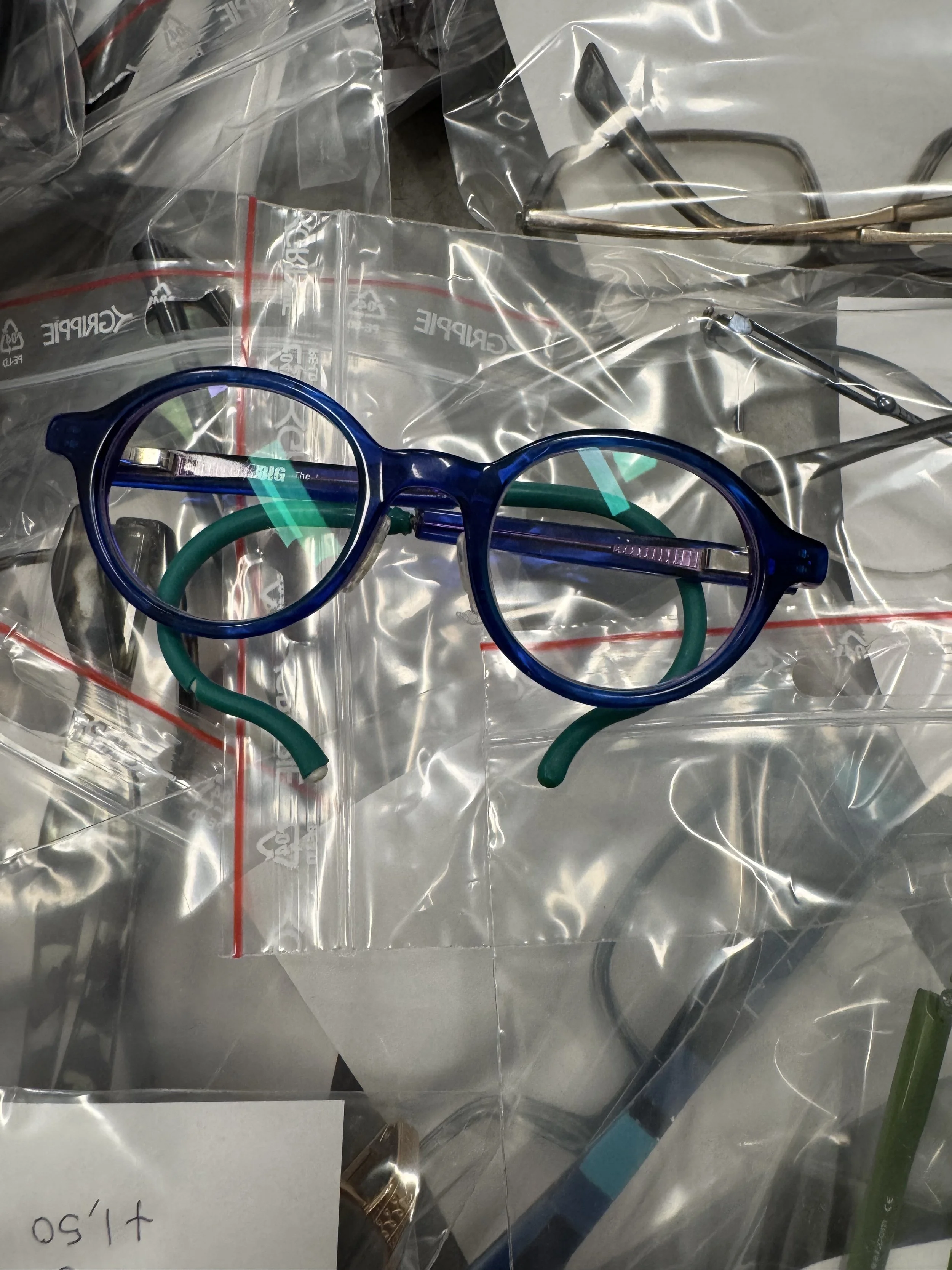A pair of blue eyeglasses with green nose pads resting on a clear plastic bag surrounded by other medical instruments and accessories.