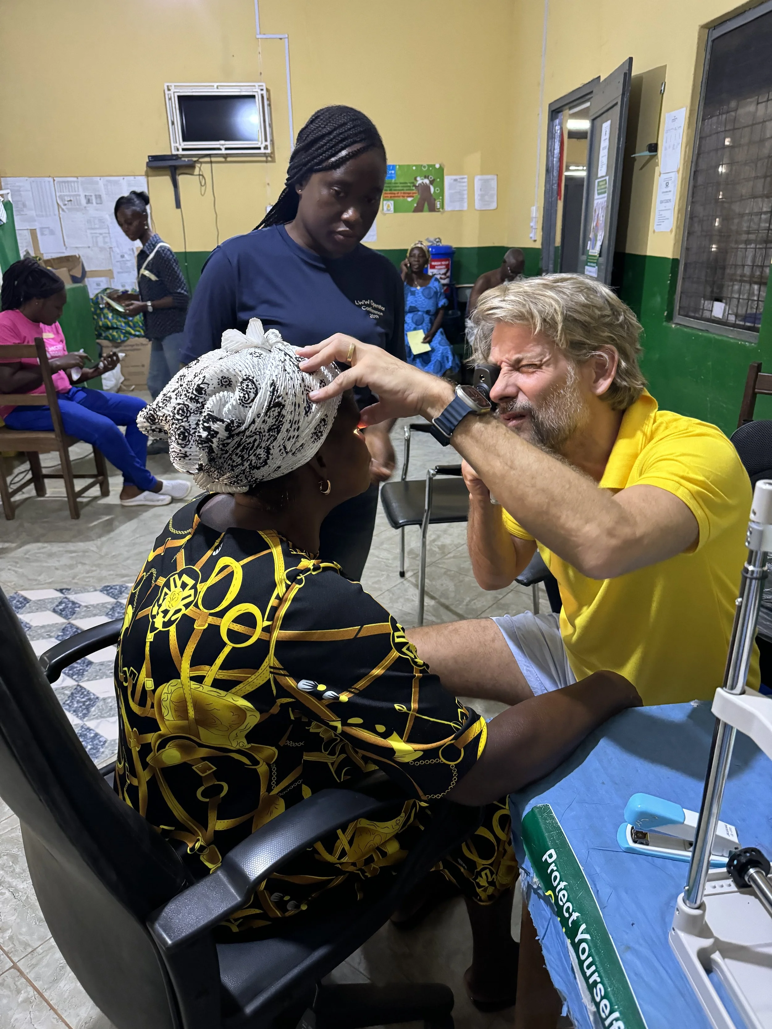 A healthcare worker examining a woman using a specialized device in a medical setting. Several people are seated in the background, some reading and others waiting, with a yellow and green interior.