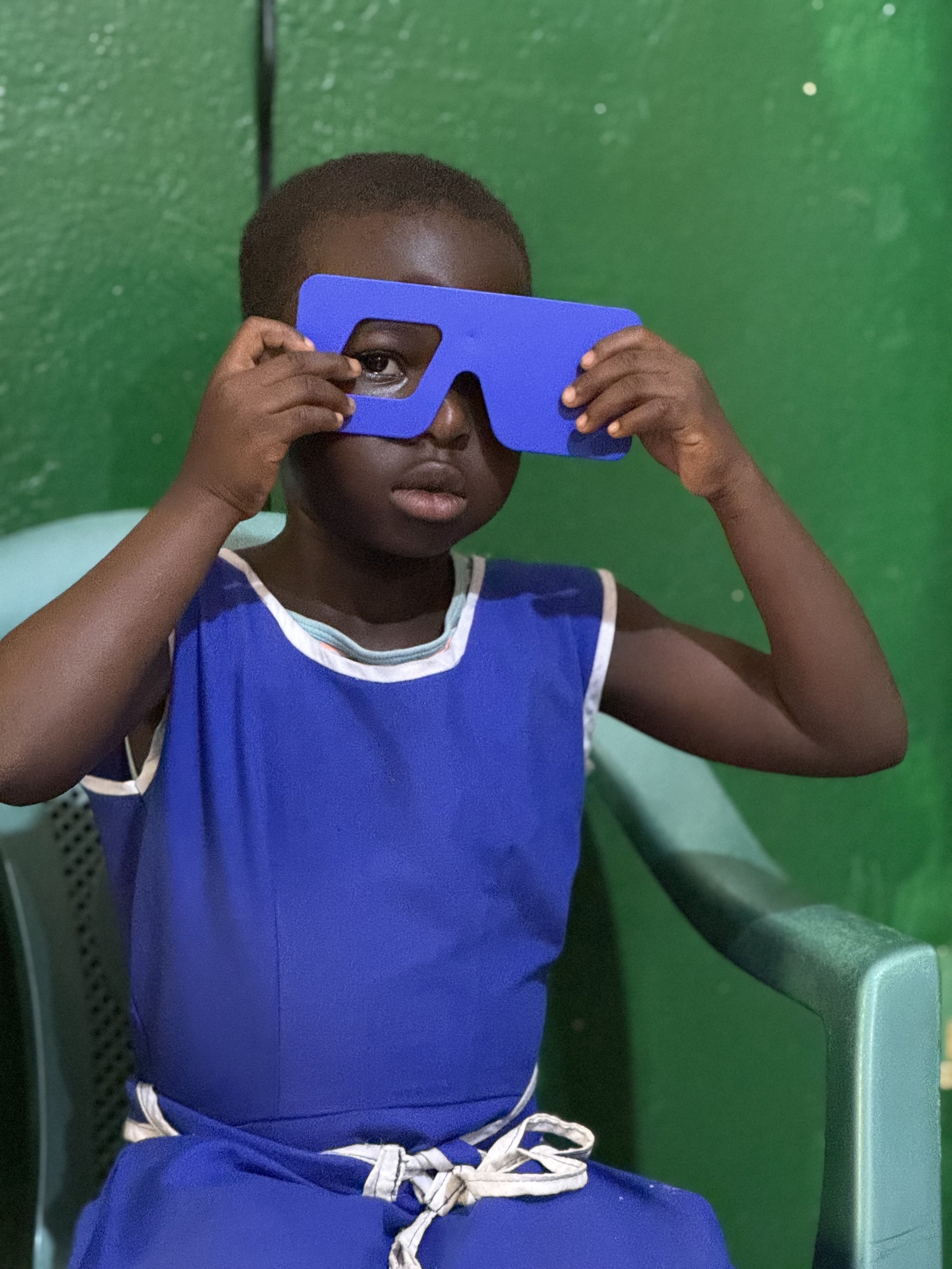 A young boy in a blue sleeveless shirt and matching pants adjusting a pair of blue glasses while seated on a green plastic chair against a green wall.