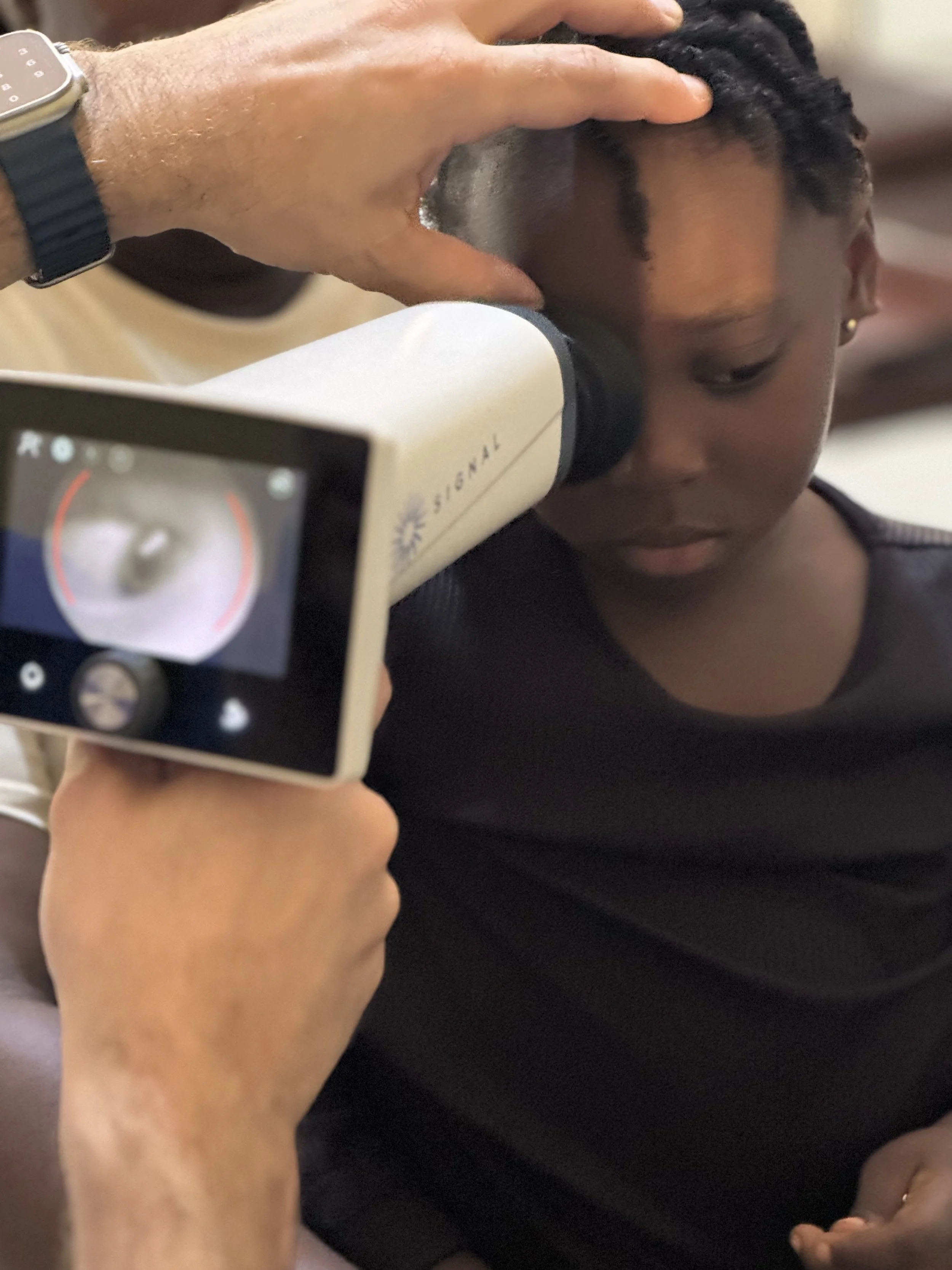 Person having eye examined with a handheld ophthalmic device, and a healthcare professional adjusting an instrument near the person's eye.