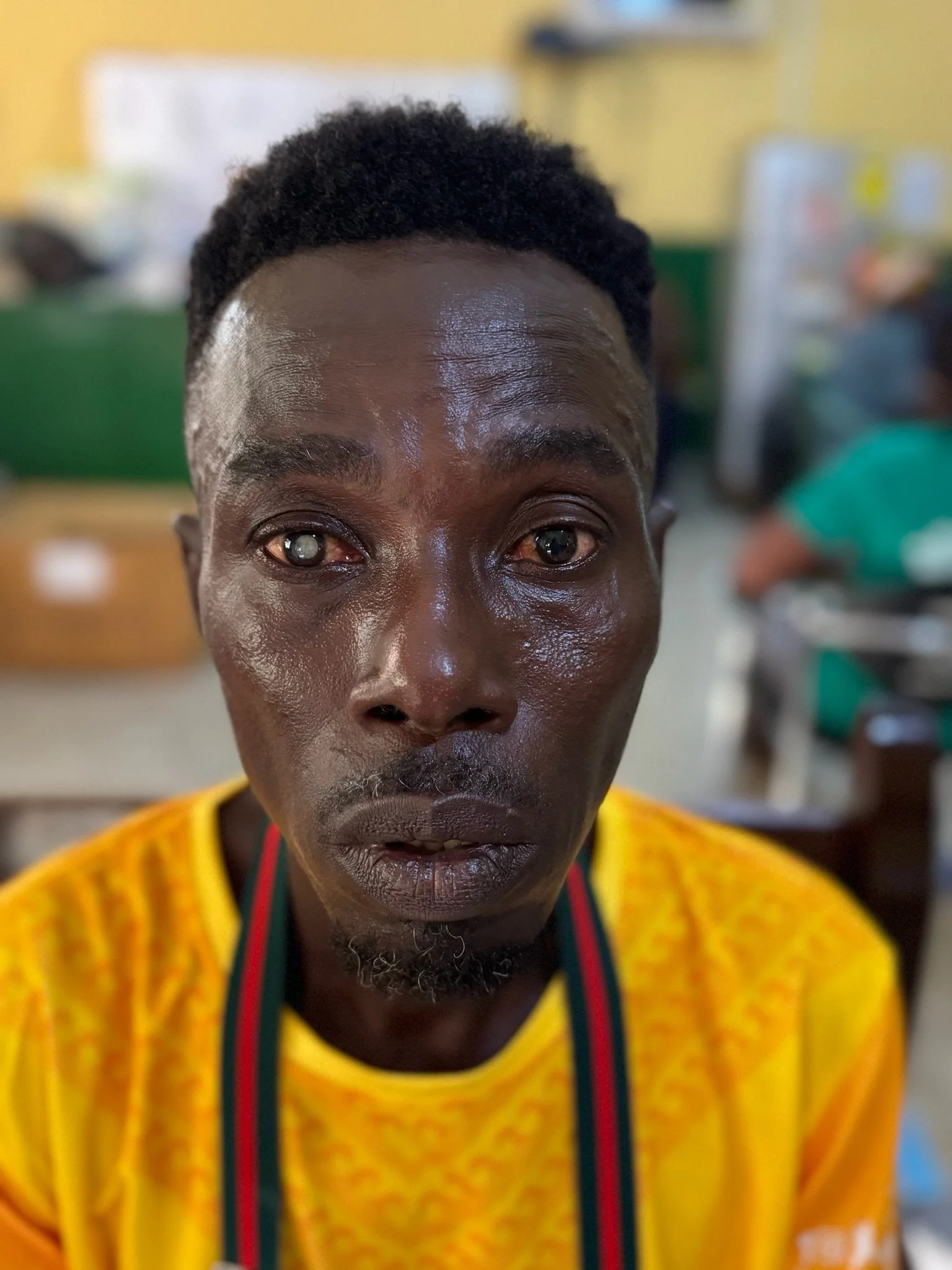 Close-up of a man with dark skin, short curly hair, wearing a yellow shirt, in a blurred indoor setting.