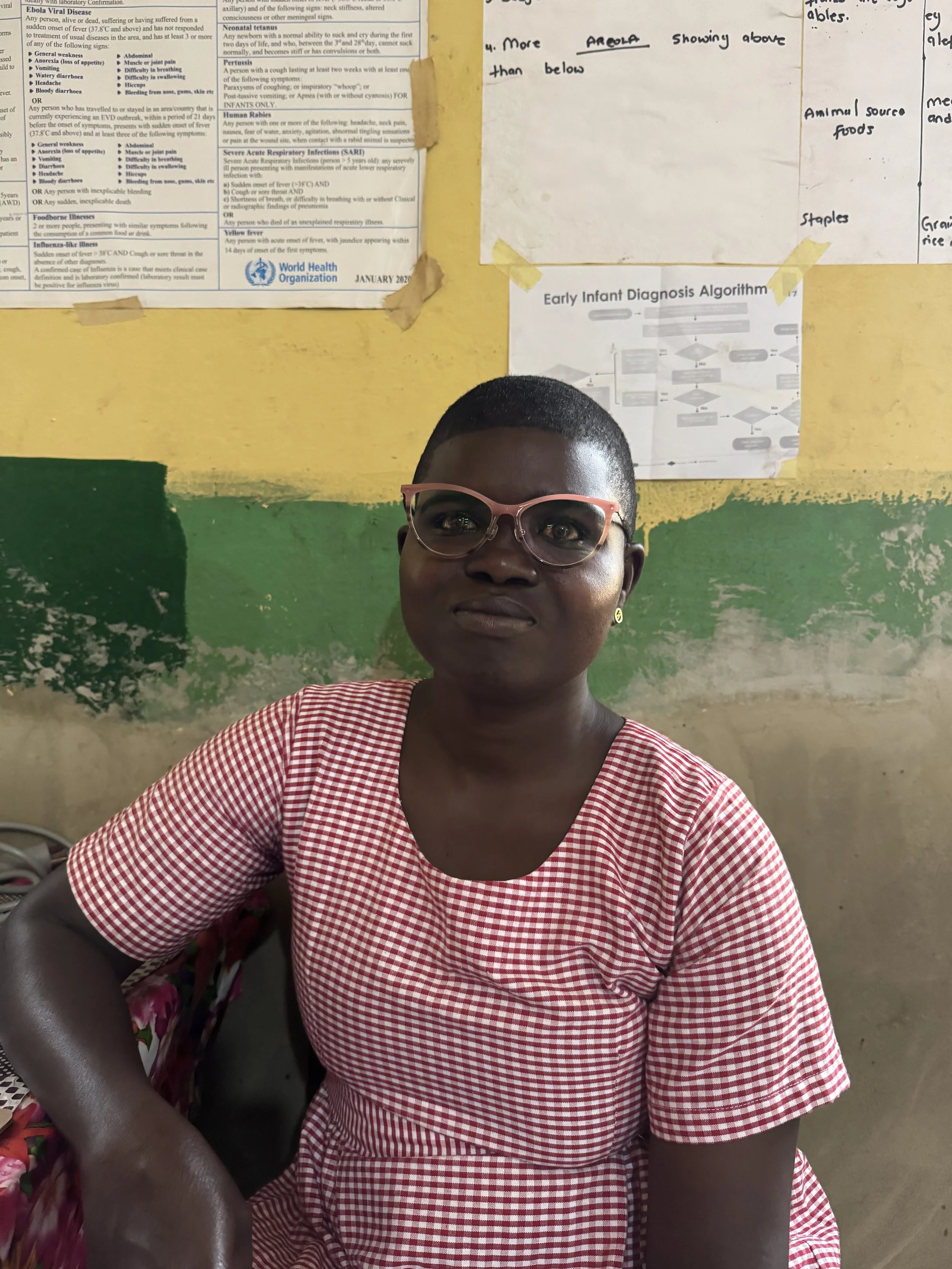 A woman with short hair, wearing glasses and a red and white checkered dress, sits in front of a wall with medical posters and a yellow and green painted background.