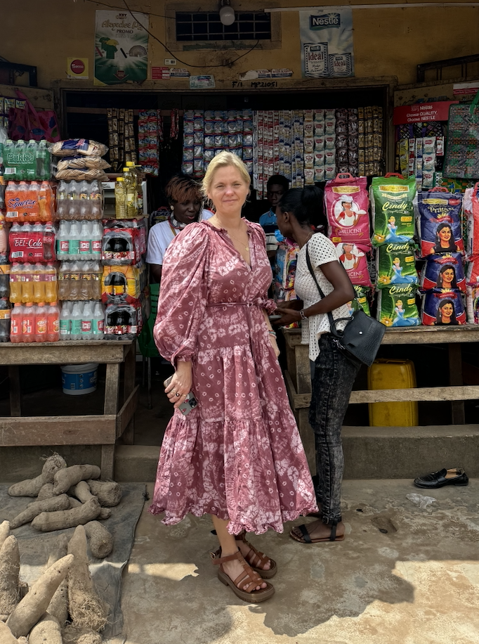 Woman in a pink dress standing at a small shop with various snacks and drinks, with other customers and shopkeepers behind her.