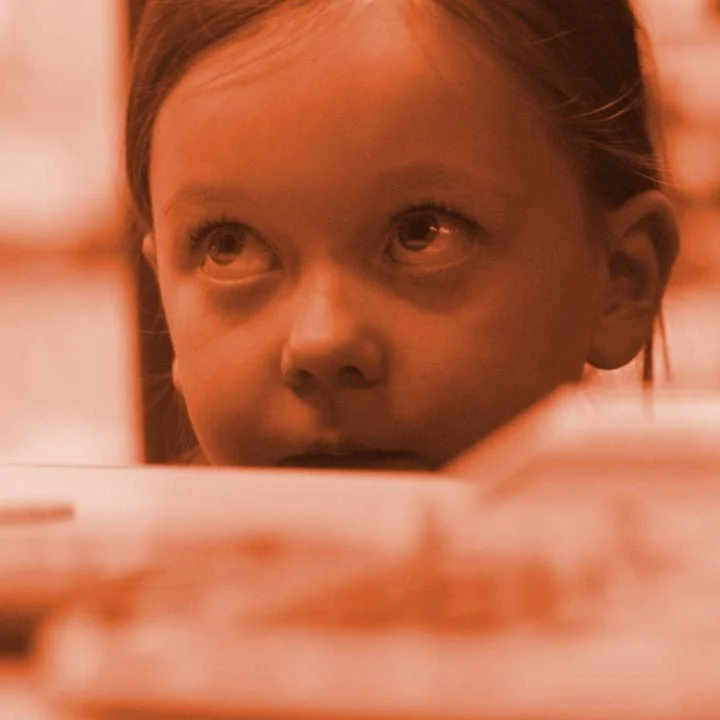 Close-up of a young girl with an inquisitive expression, looking upward, with a blurred background in warm lighting.