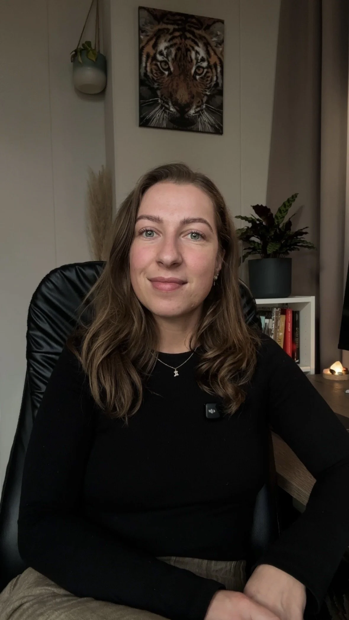 Een vrouw met bruin, golvend haar zit aan een bureau in een kamer met boekenkast en planten. Op de muur hangt een schilderij van een tijger.
