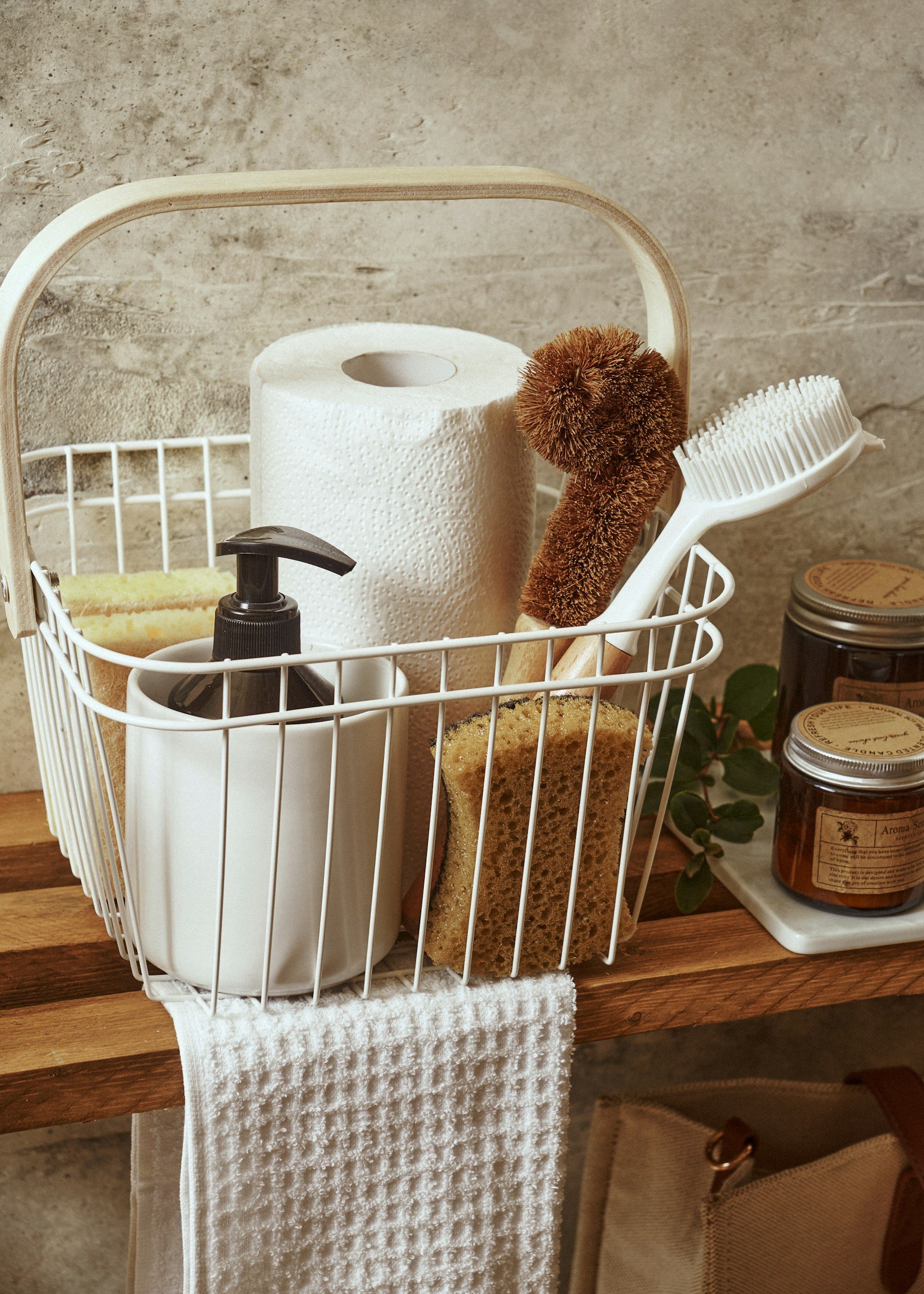 A white wire basket on a wooden surface holding a paper towel roll, a sponge, a bottle of liquid soap, a brown scrub brush, and a kitchen sponge. To the right, there are two jars on a small tray with green foliage next to them.