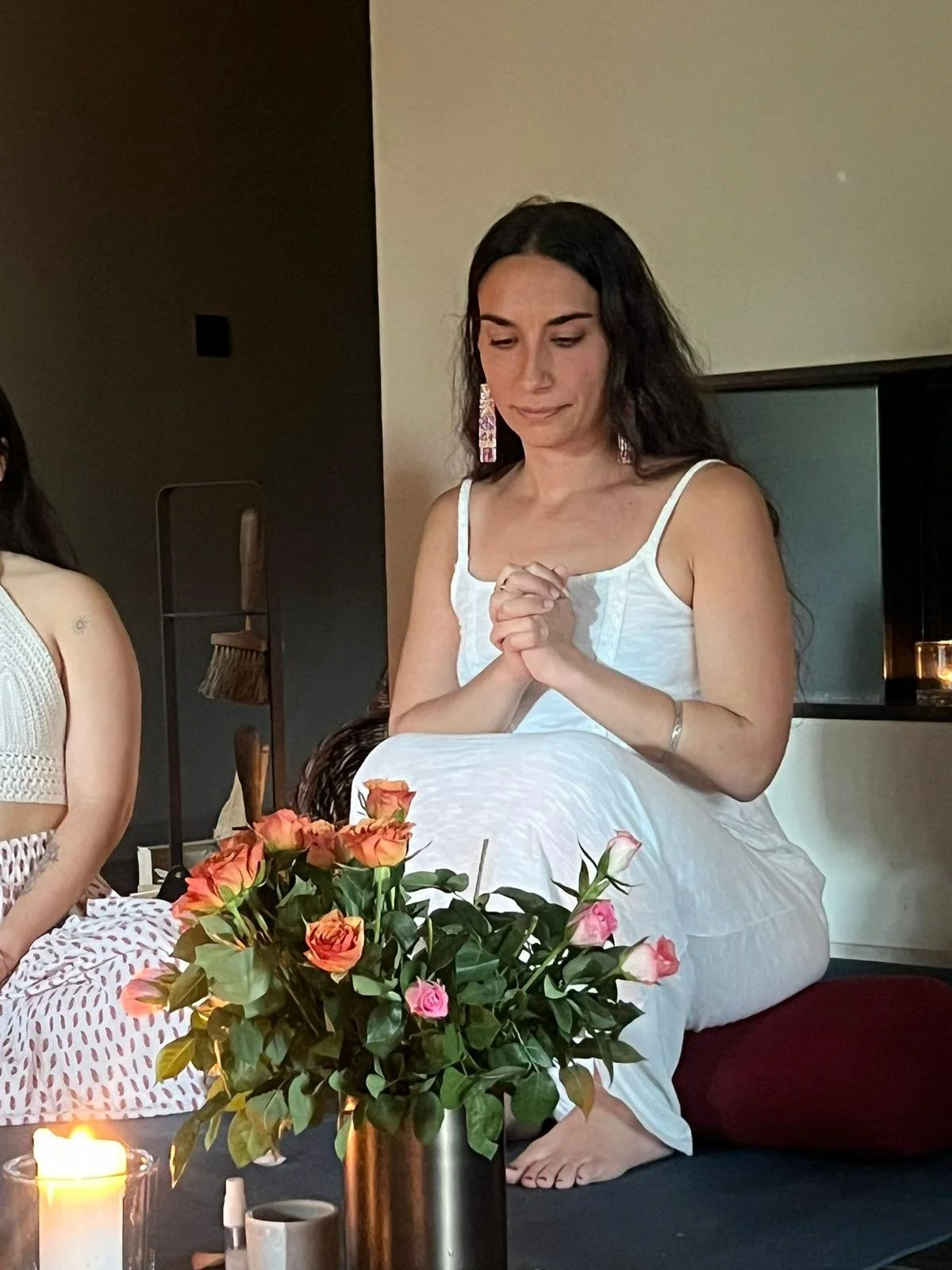 Une femme en robe blanche, assise avec les mains jointes et les yeux fermés, près d'un bouquet de roses colorées, dans un intérieur faiblement éclairé.