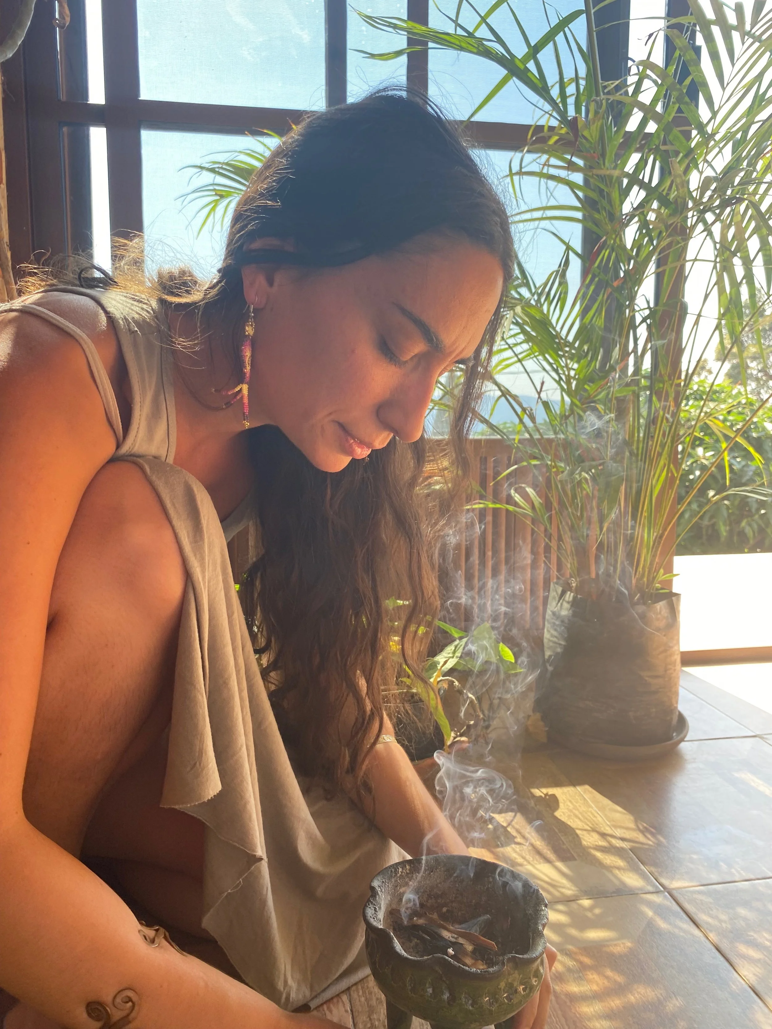 Une femme assise à une table, tenant un petit récipient en pierre ornementale, avec de la fumée qui en sort. Elle porte une robe beige et des boucles d'oreilles colorées, avec un tatouage au poignet. Il y a une grande plante verte en arrière-plan près d'une fenêtre en journée.