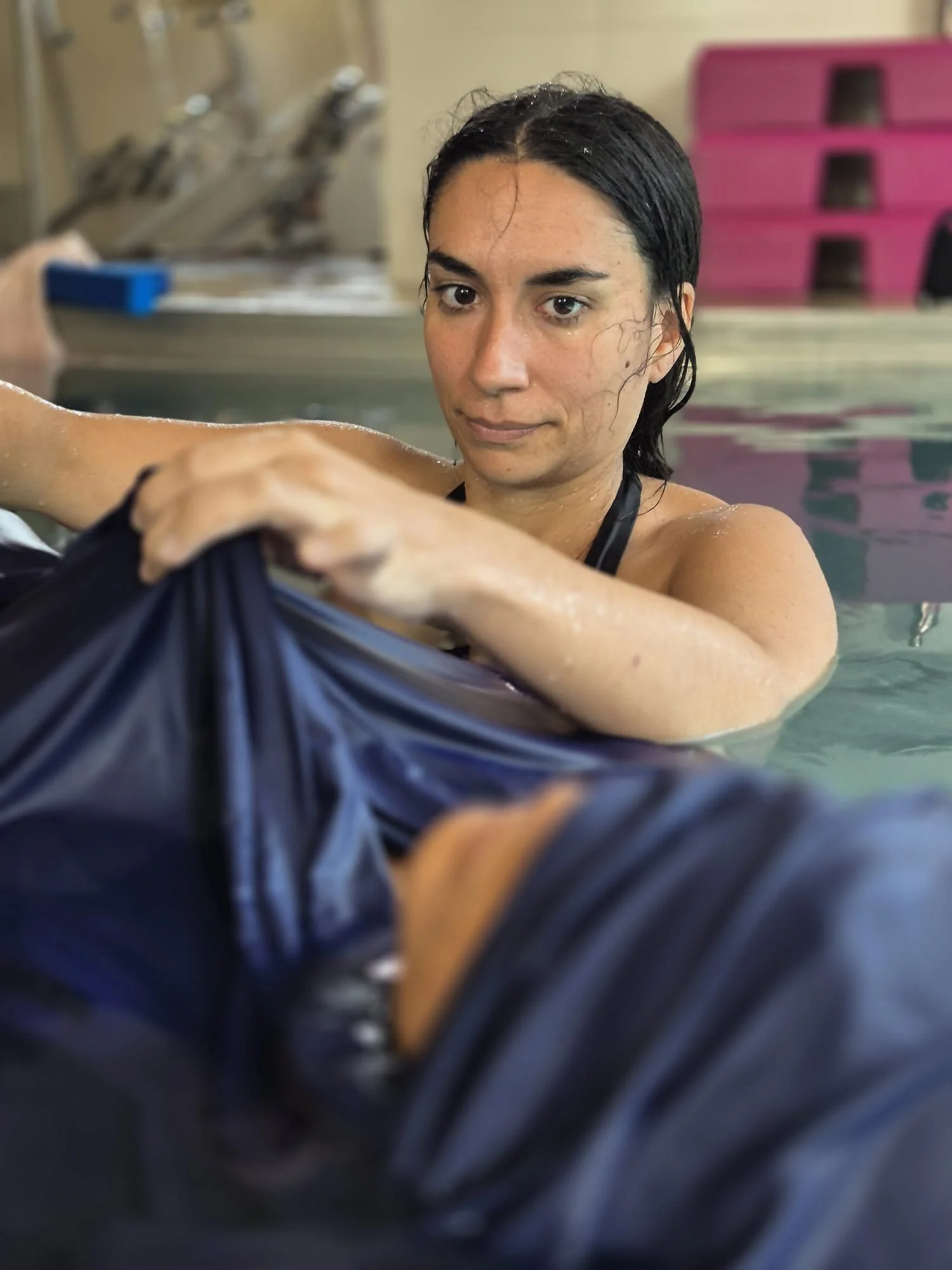 Une femme nage dans une piscine intérieure, elle tire sur une tenue ou un vêtement de couleur foncée, avec des cheveux mouillés et un regard concentré.