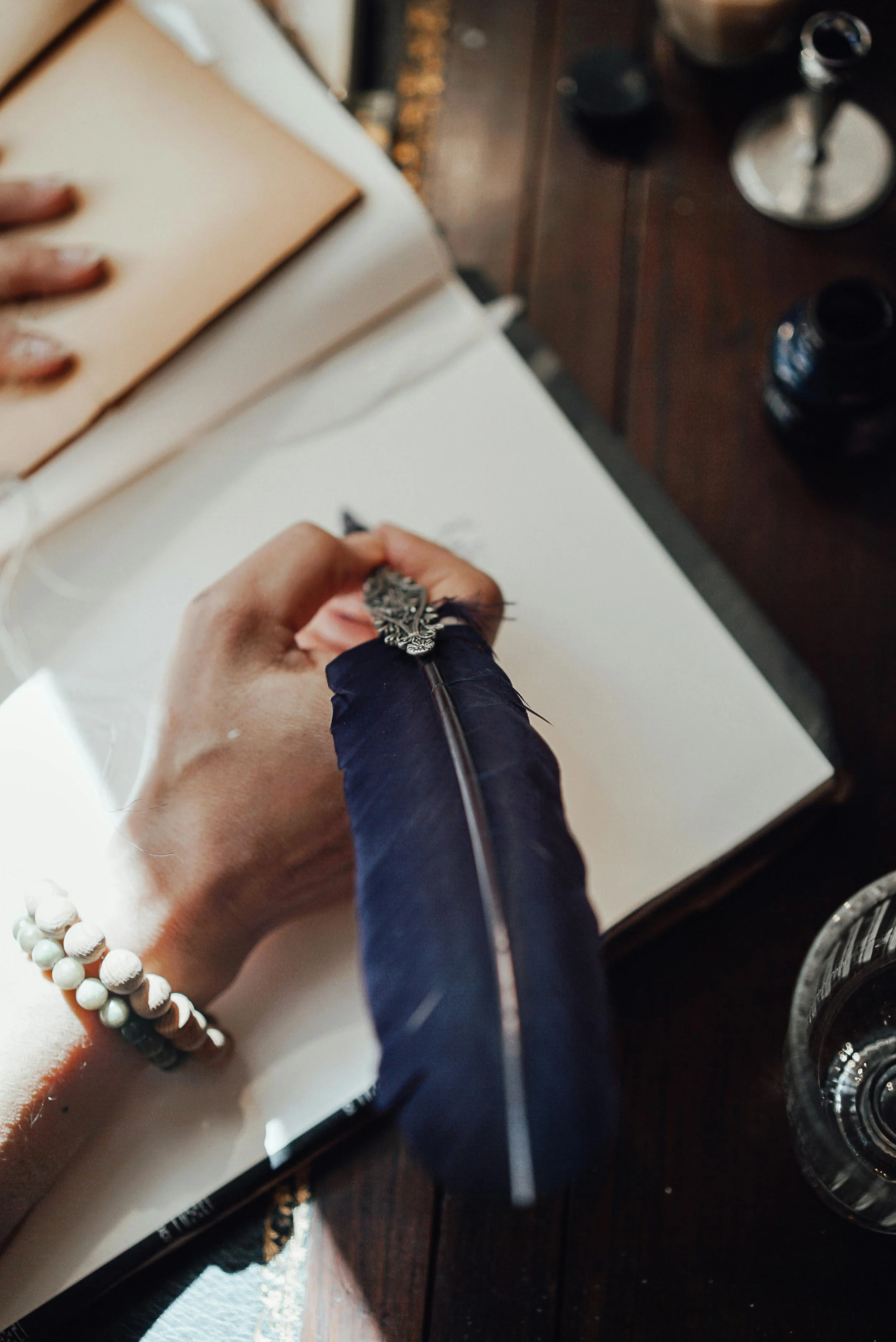 Une personne écrit dans un carnet blanc avec un stylo à plume décoré, portant un bracelet de perles, à une table en bois avec plusieurs fioles de vernis à ongles et une liseuse.