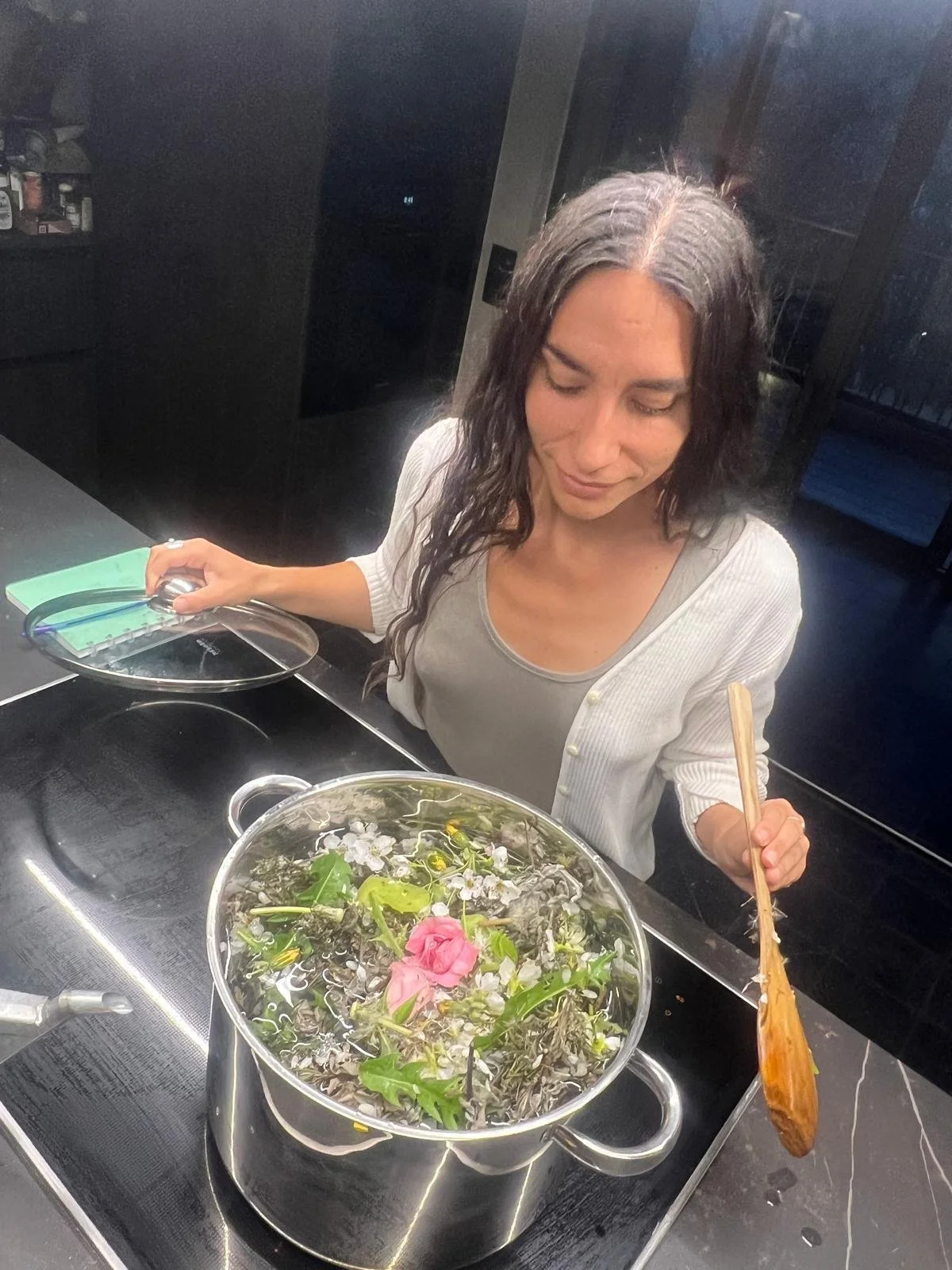 Une femme regarde un bol rempli de fleurs et de feuilles dans une cuisine, tenant une cuillère en bois. Elle prépare un hydrolat de rose pour la cérémonie de la rose pour son cercle de femmes.