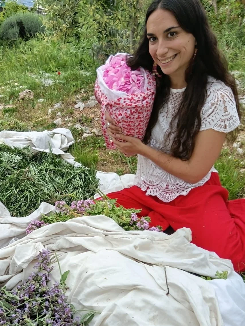 Femme souriante tenant un bouquet de fleurs roses, assise dans une nature avec des plantes et des fleurs autour, portant un vêtement blanc en dentelle et un pantalon rouge.
