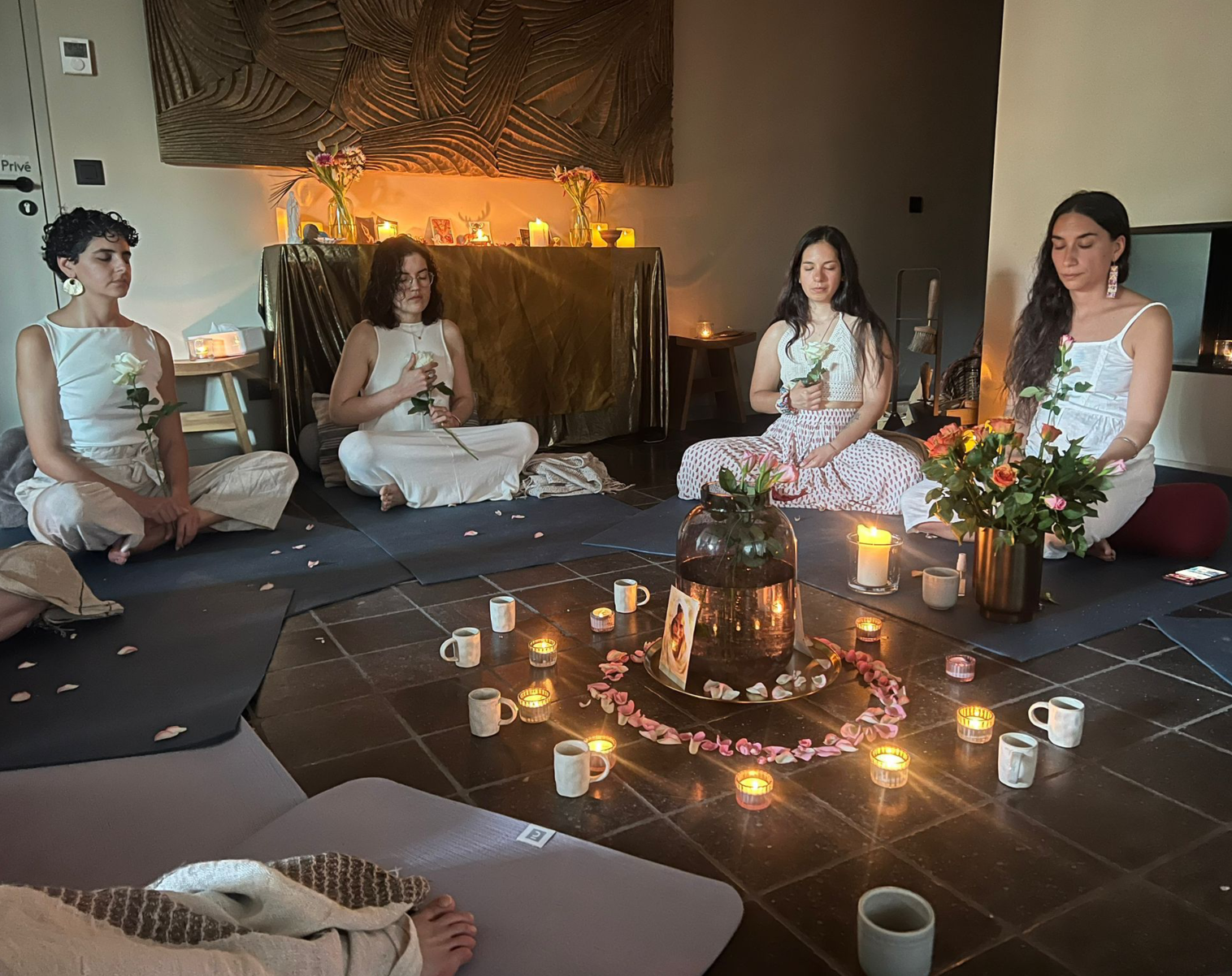 Quatre femmes assises en méditation dans une pièce, entourées de bougies, de fleurs, et de décorations, dans une ambiance calme et spirituelle.