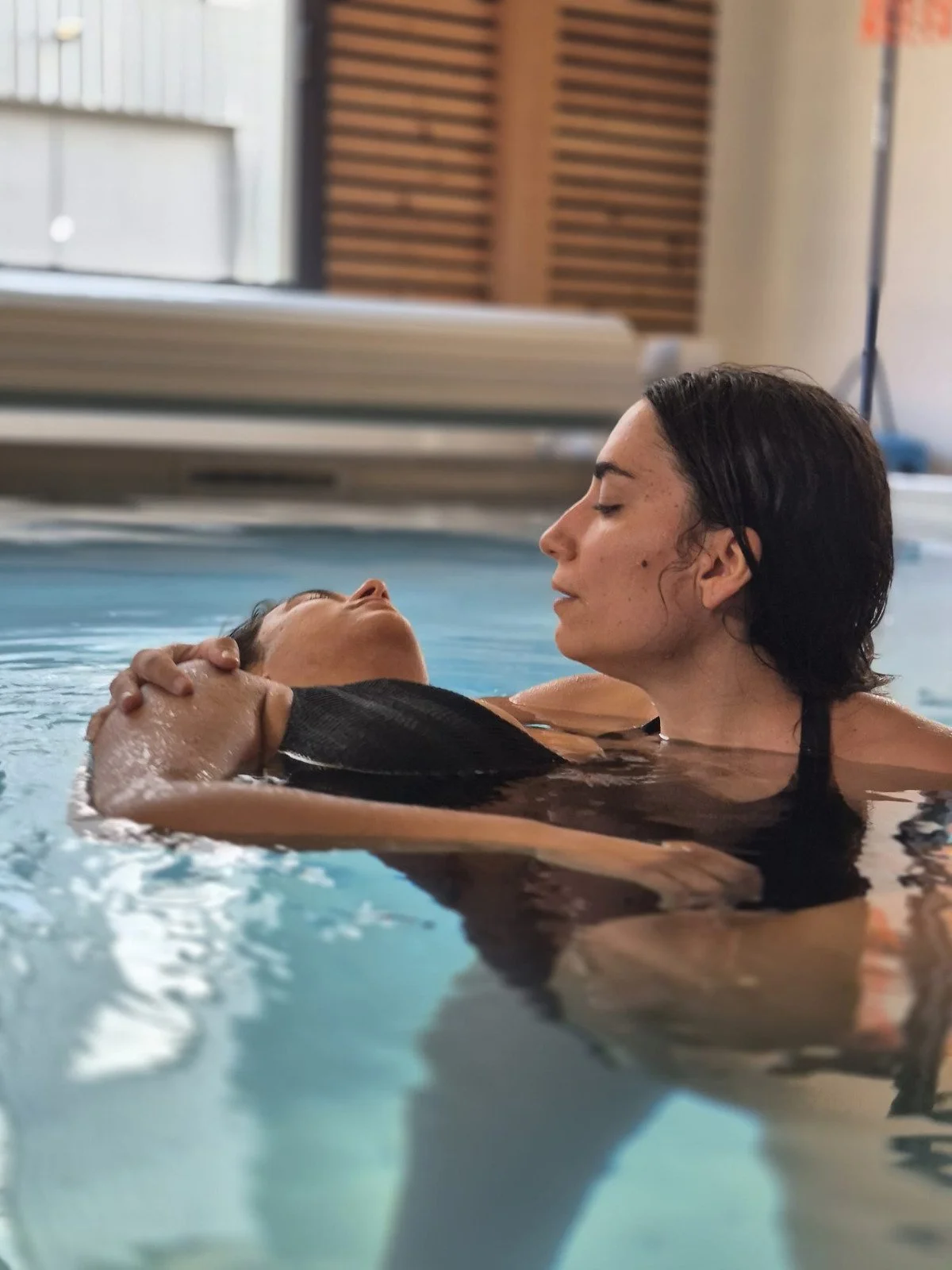 Deux femmes dans une piscine, l'une d'elles aidant l'autre à flotter, en intérieur.