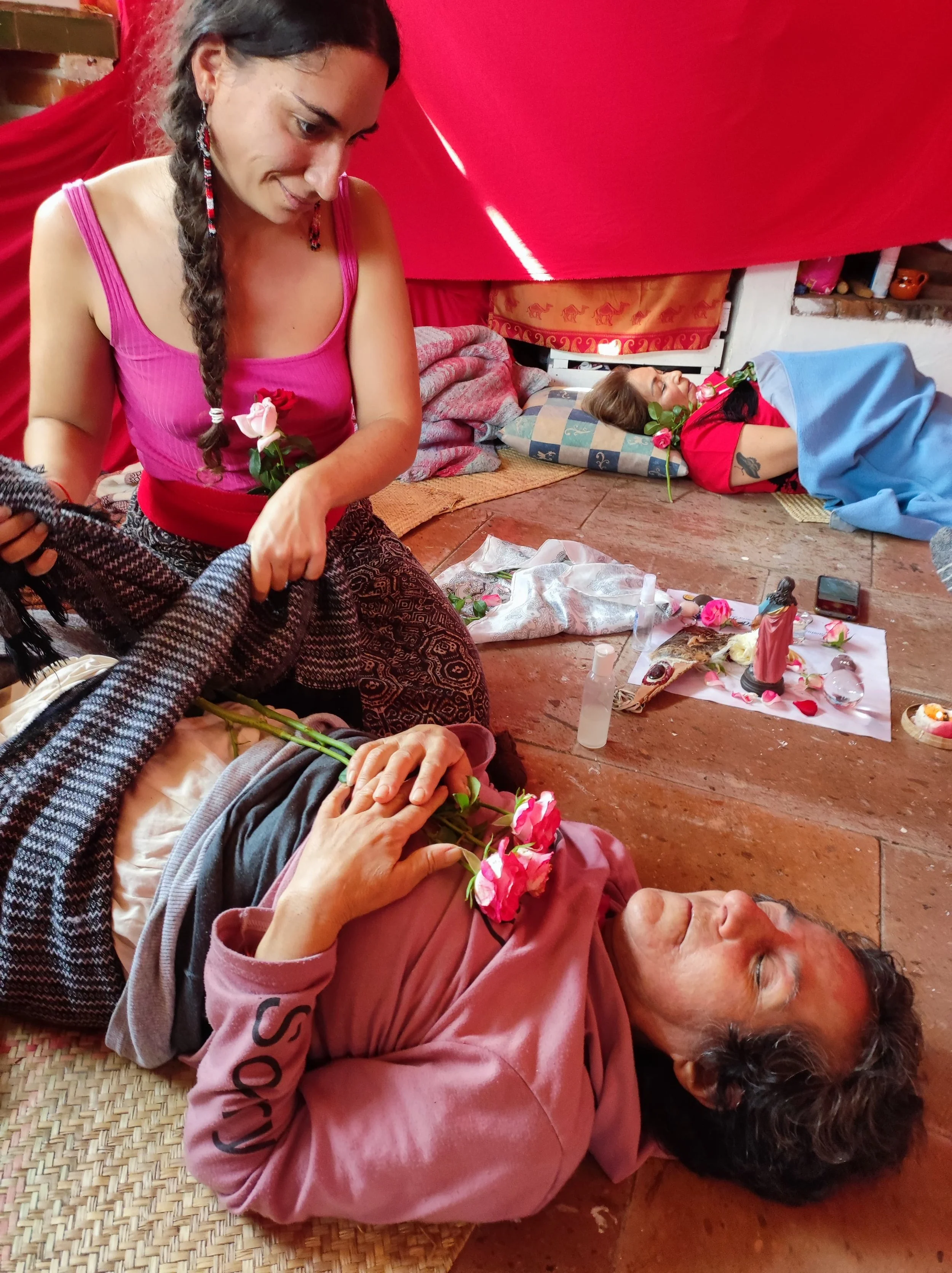Une femme allongée sur le sol, avec des fleurs roses sur elle, lui faisant un soin rebozo, aidé par les écharpes mexicaines qui accompagnent lors des bouleversements de la vie;